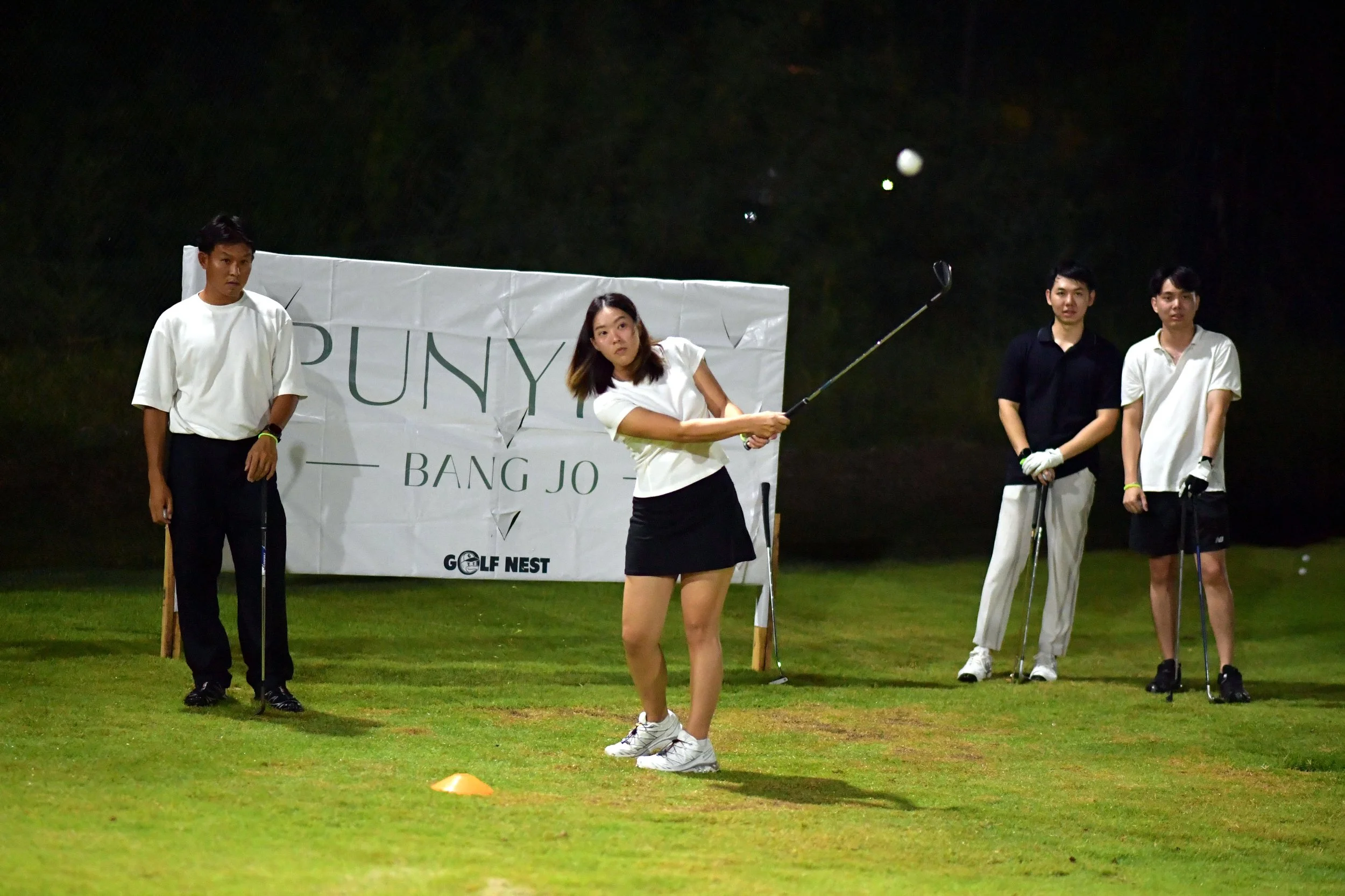A woman playing golf on a night course with four other people watching. She is mid-swing, holding a golf club, on a grass field with a sign in the background.