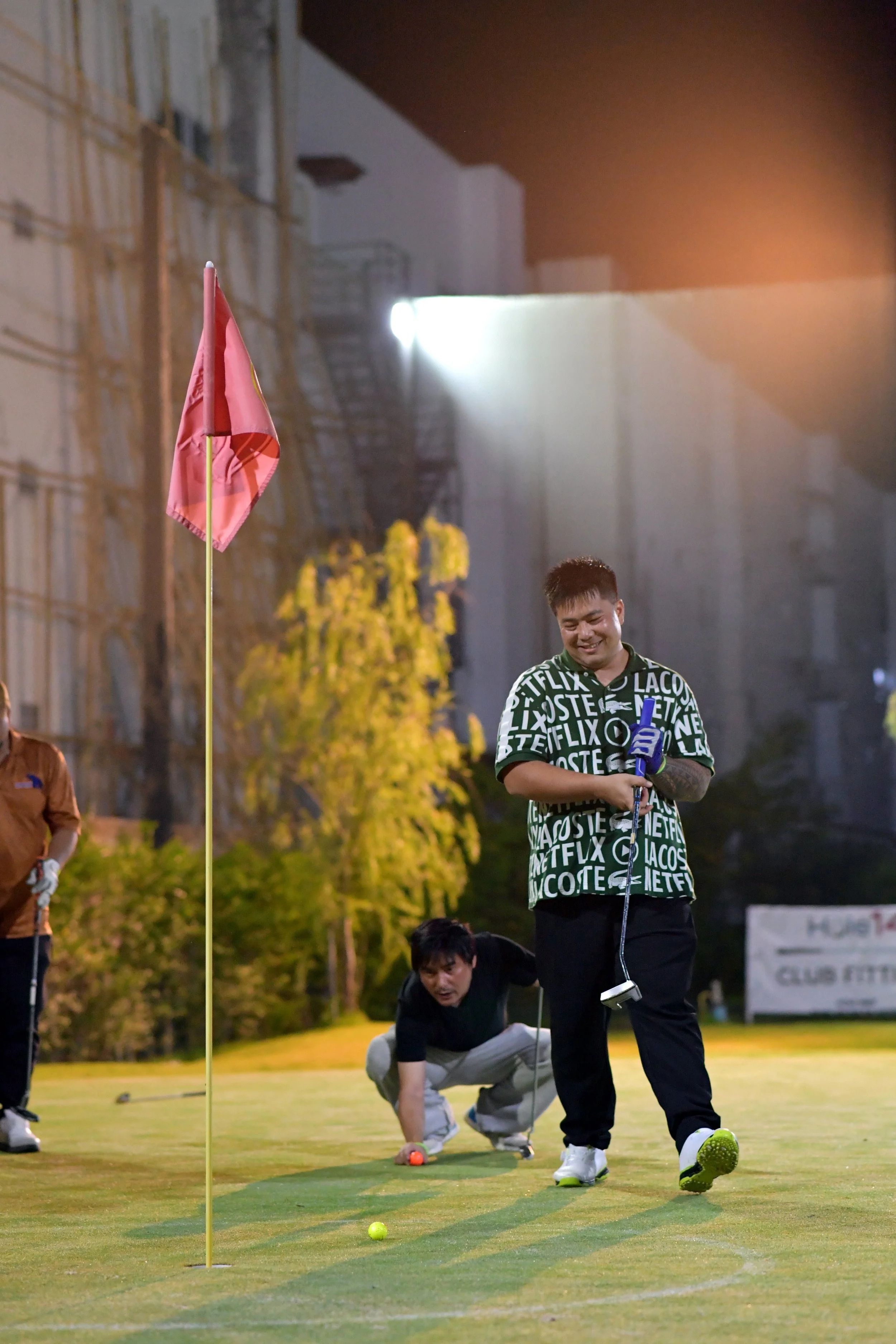 Group of people playing mini-golf outdoors at night. One person is smiling and holding a golf putter, with others preparing to putt or watching nearby. The scene is illuminated by outdoor lighting, with trees, a flag, and a building visible in the ba