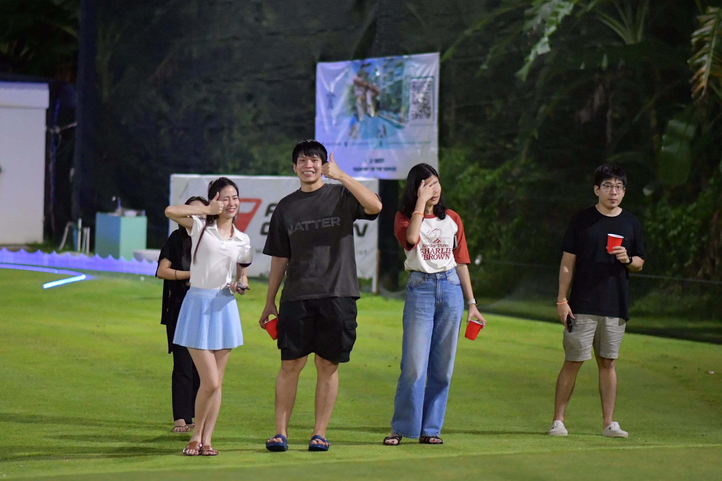 Group of five young people standing on a green field at night, some are smiling and giving thumbs up, others are covering their faces or holding red cups.