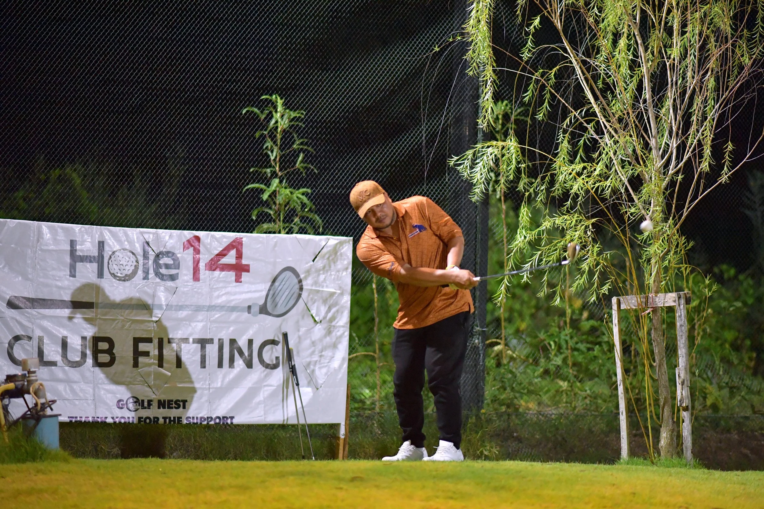 A man in an orange shirt and cap playing golf on a green, nighttime setting. There is a banner with text 'Hole 14' and 'Club Fitting' in the background.