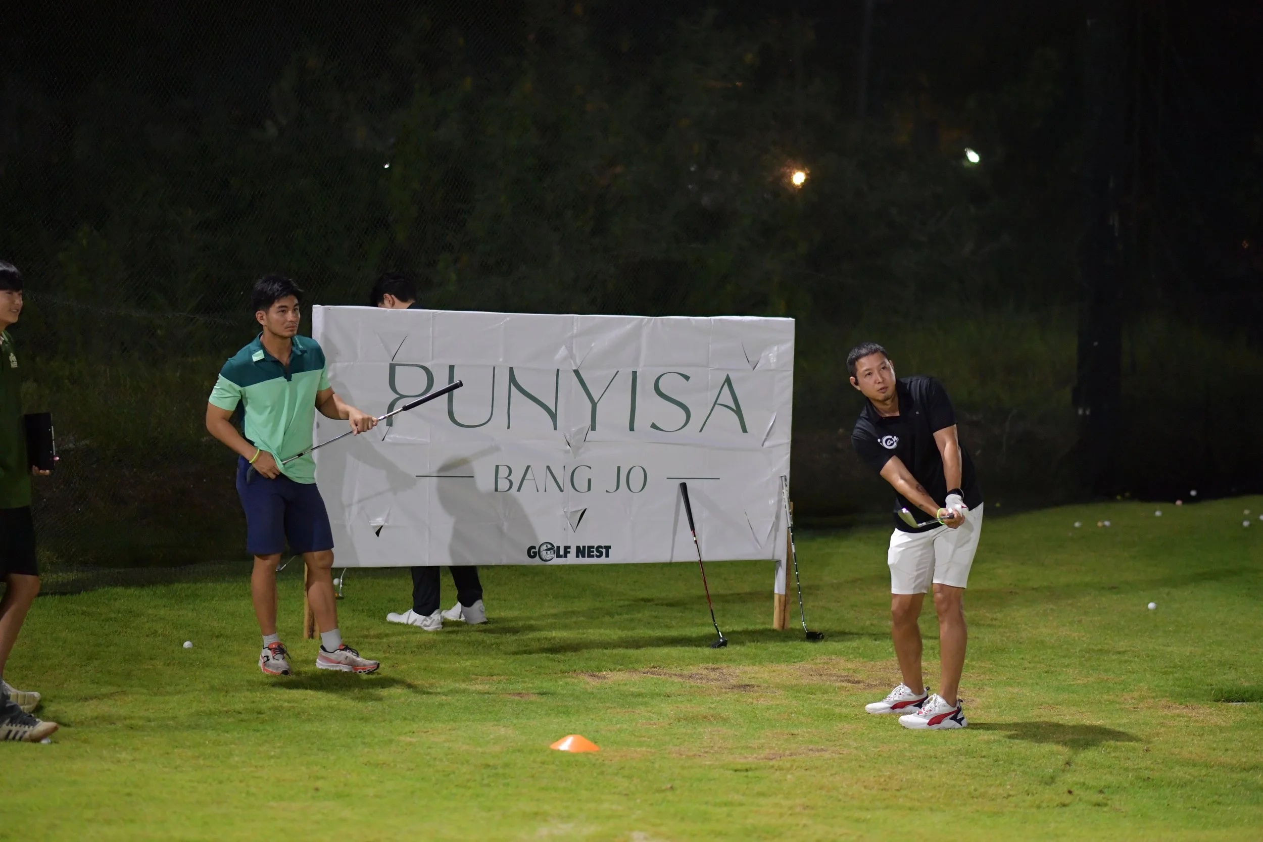 A man in a black shirt and white shorts swings a golf club at a golf tee, with a golf ball on the grass. Other people stand nearby, and a large banner reads "PUNYISA BANG JO GOLF NEST." The scene is illuminated at night on a golf course.