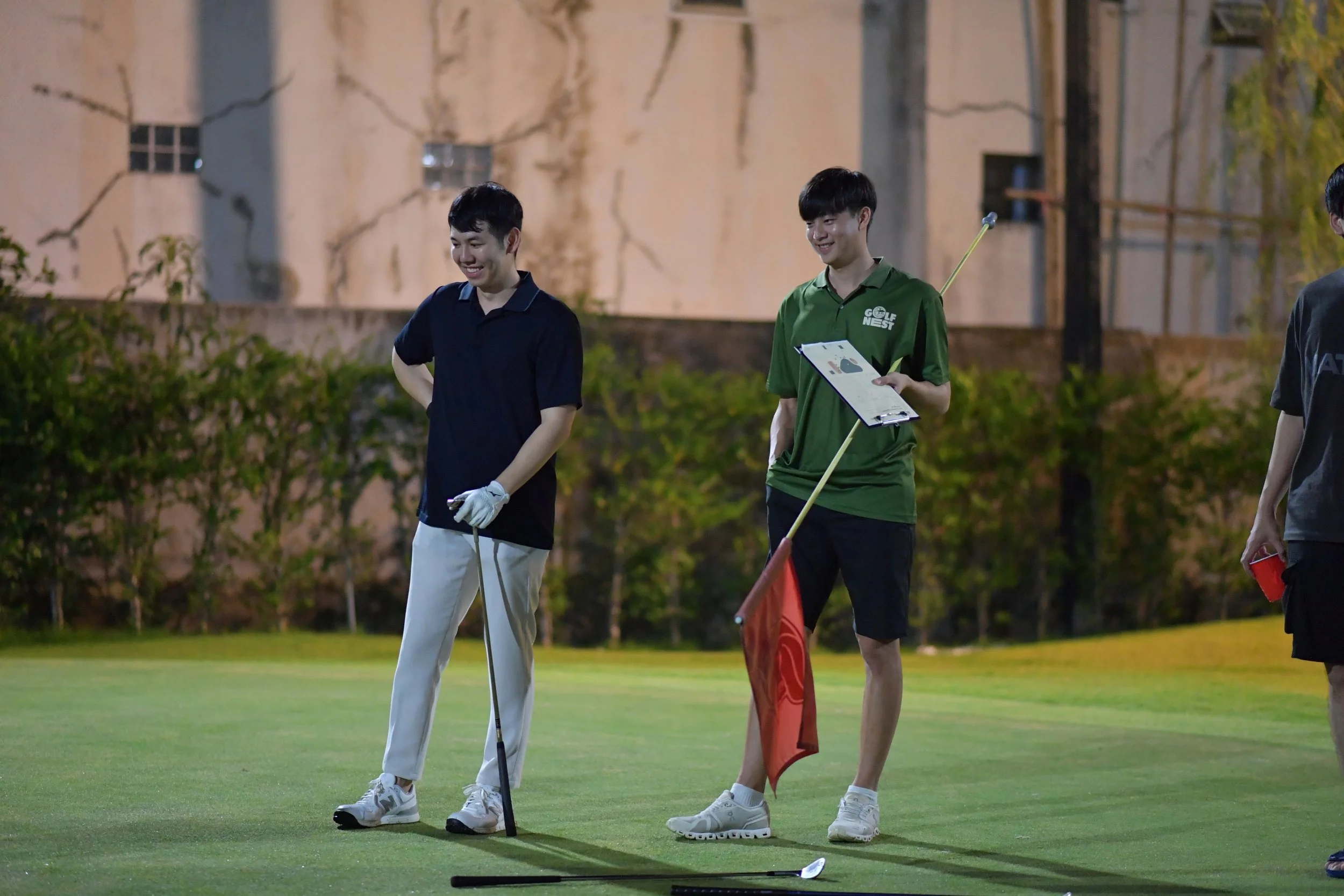 Two young men on a golf course at night, smiling, with one holding a golf club and the other holding a clipboard and a long golf flag, with a background of trees and a building.