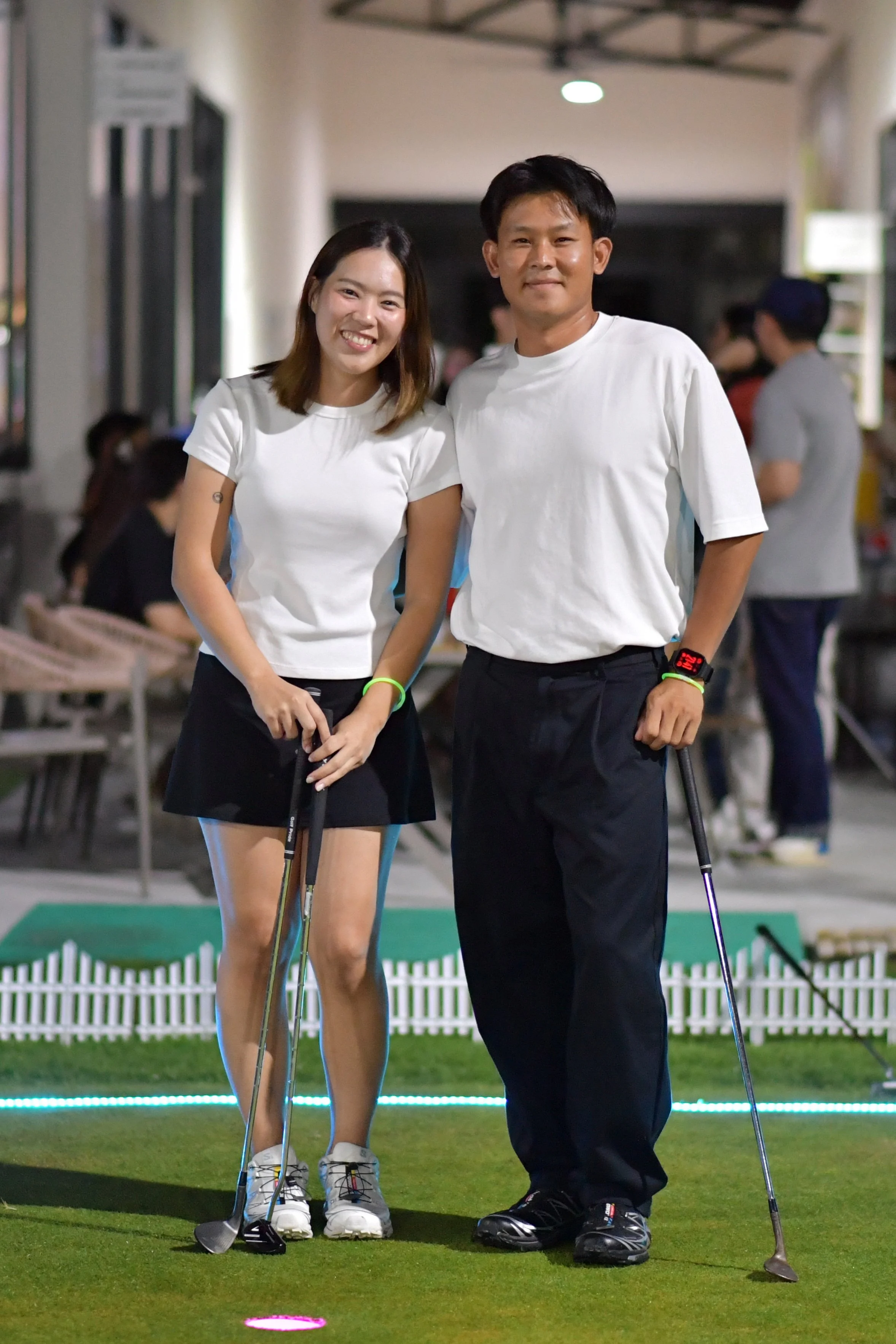 A young woman and man standing side by side on a mini-golf course at night, both holding golf clubs and smiling at the camera.