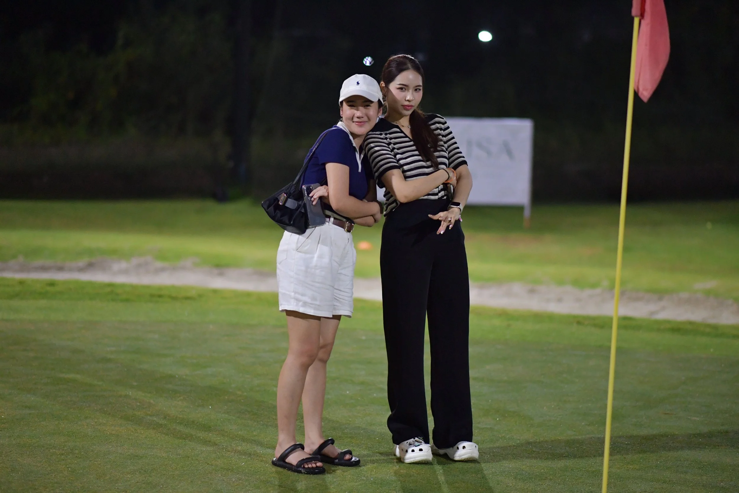 Two women standing on a golf course at night, near a golf hole with a flag, posing for the camera.