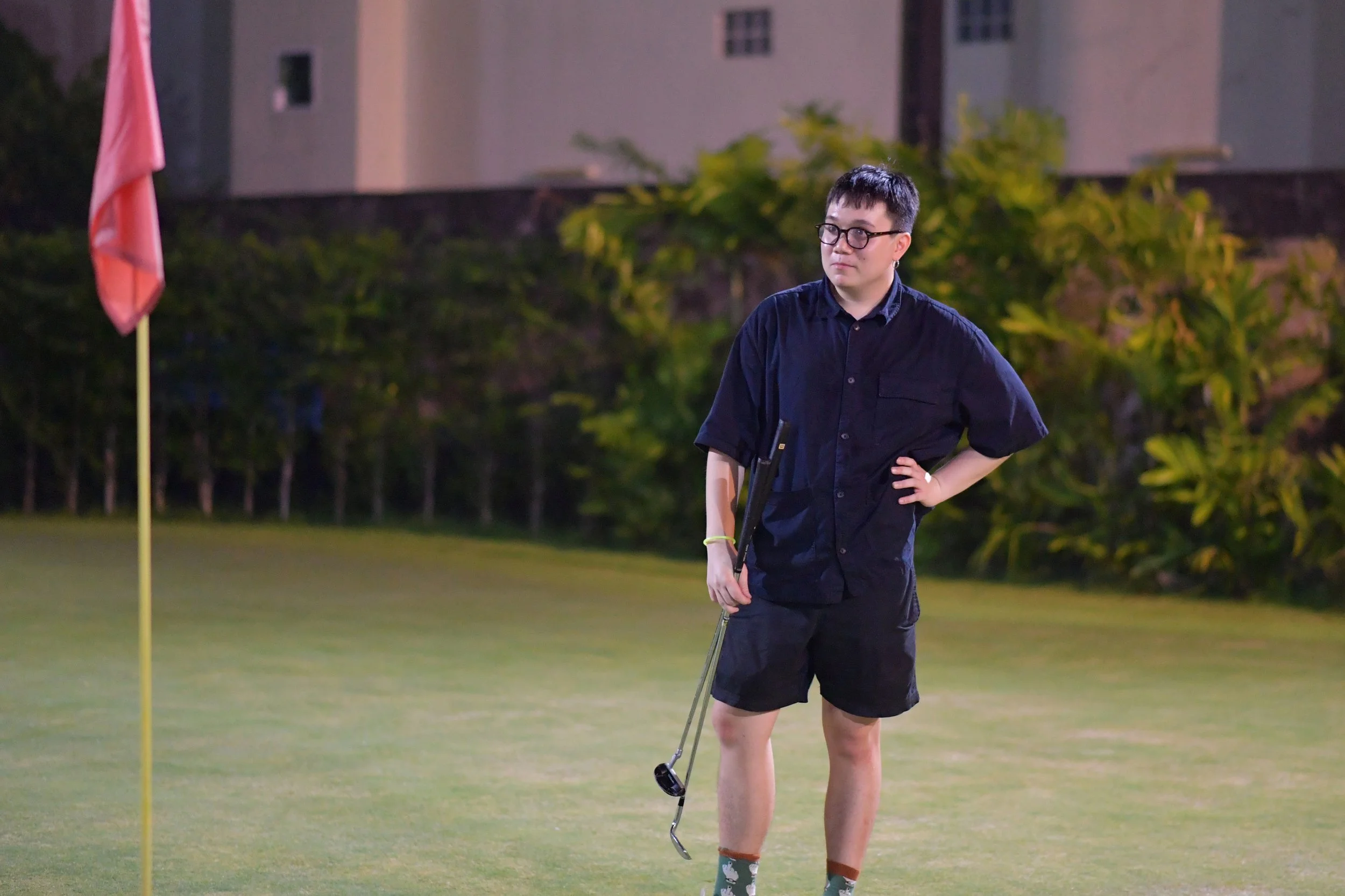 A young man standing on a golf course at night, holding a golf club, wearing a dark shirt, shorts, and glasses, with one hand on his hip and a serious expression.