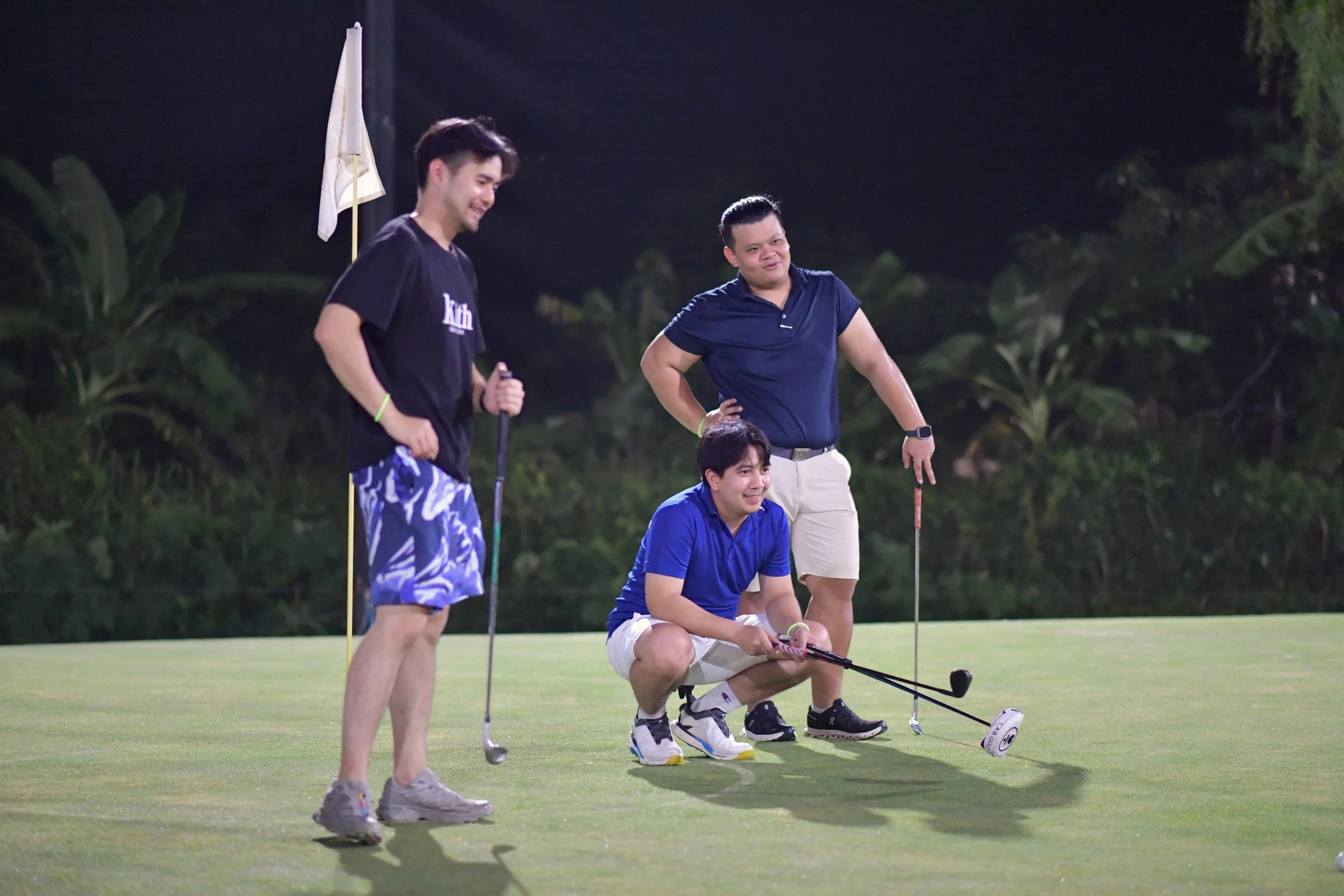 Three men playing golf on a green lawn at night, one is kneeling and two are standing, with a golf flag in the background, smiling and holding golf clubs.