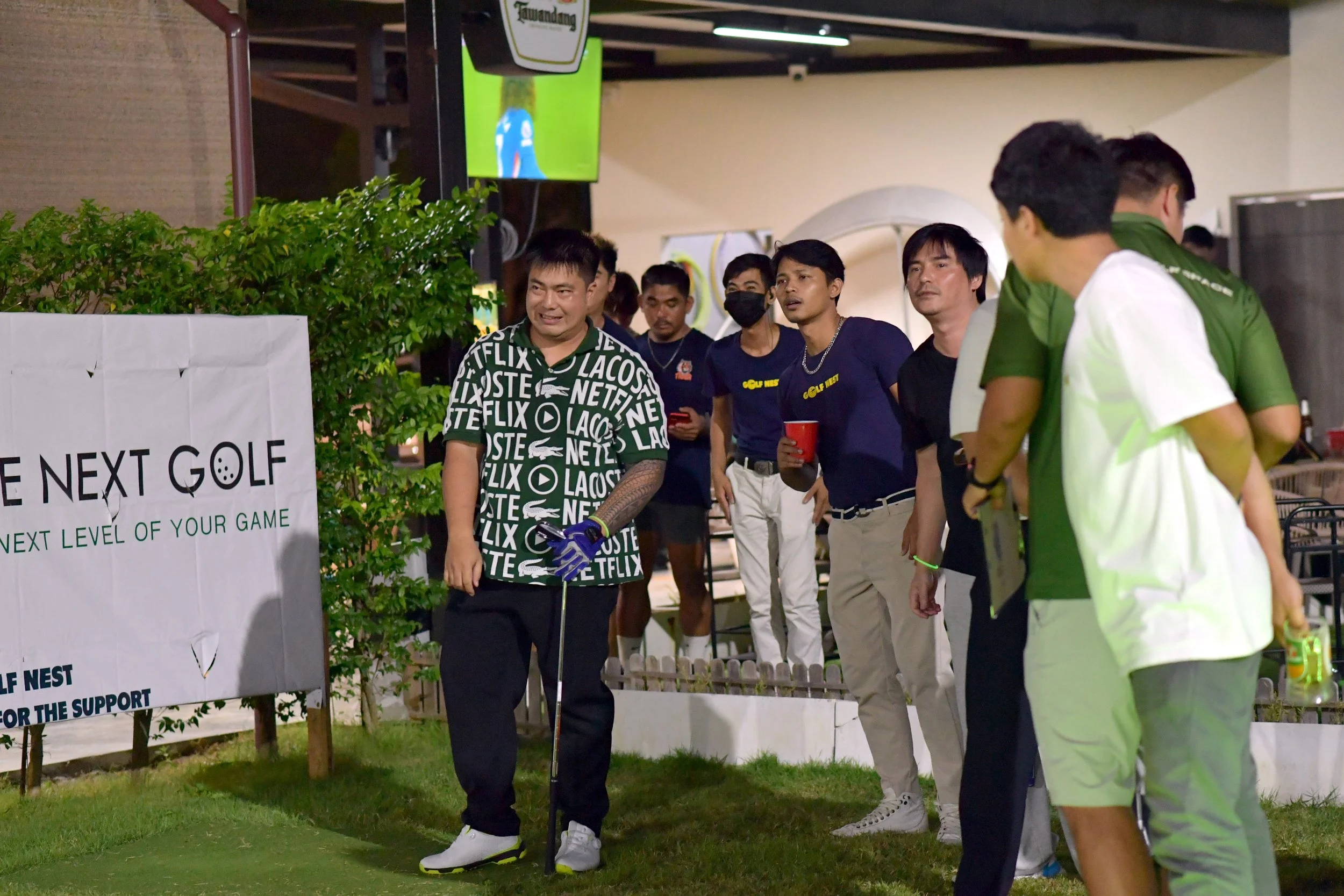 Group of people at a golf event, with a sign that reads 'The Next Golf' and a man holding a golf club, outdoors at night.