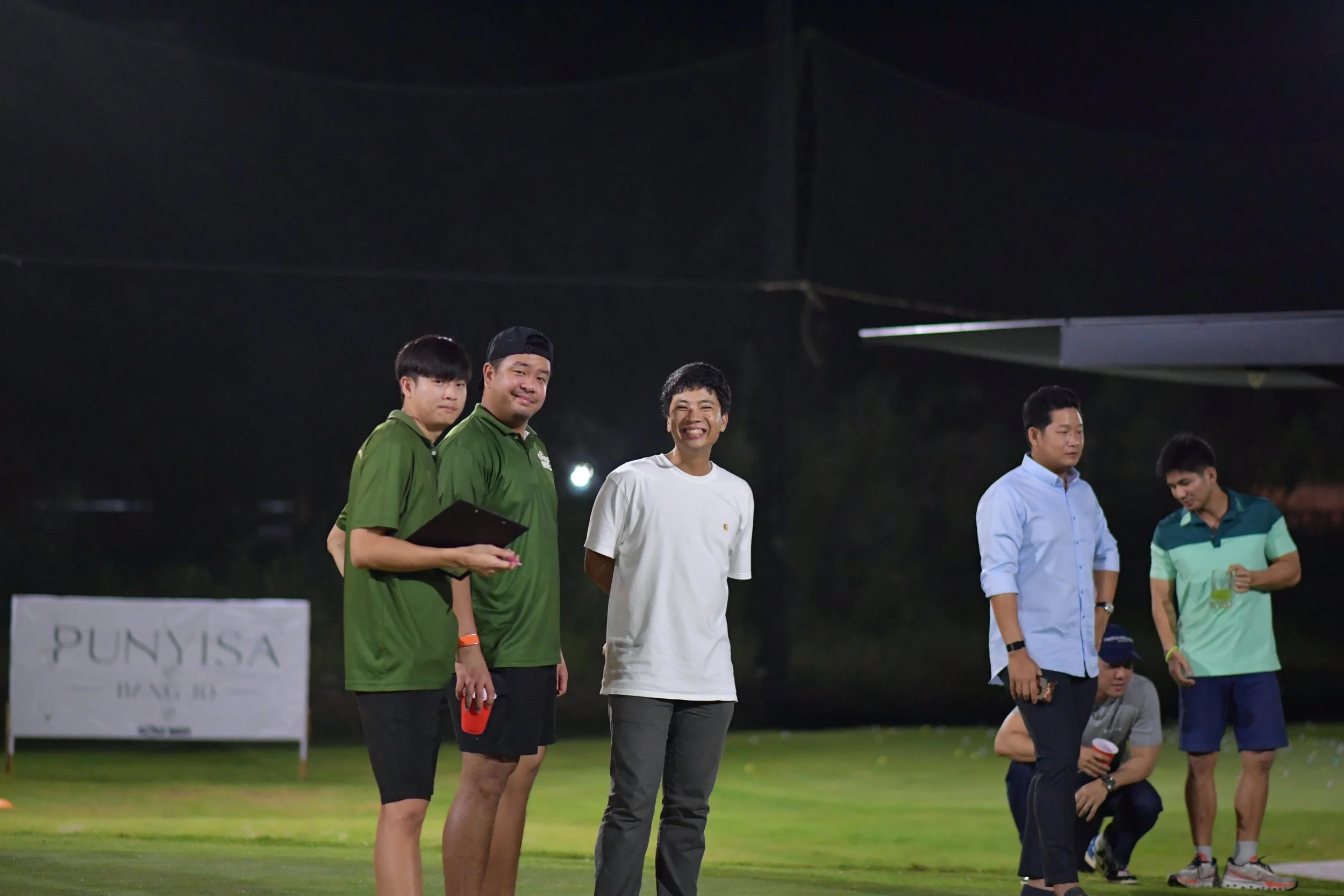 Group of five men standing on a grassy field at night, some smiling and others looking down. Two men wear green uniforms, one man in white, another in a light blue shirt, and the last in a striped green and blue shirt. A sign in the background reads 