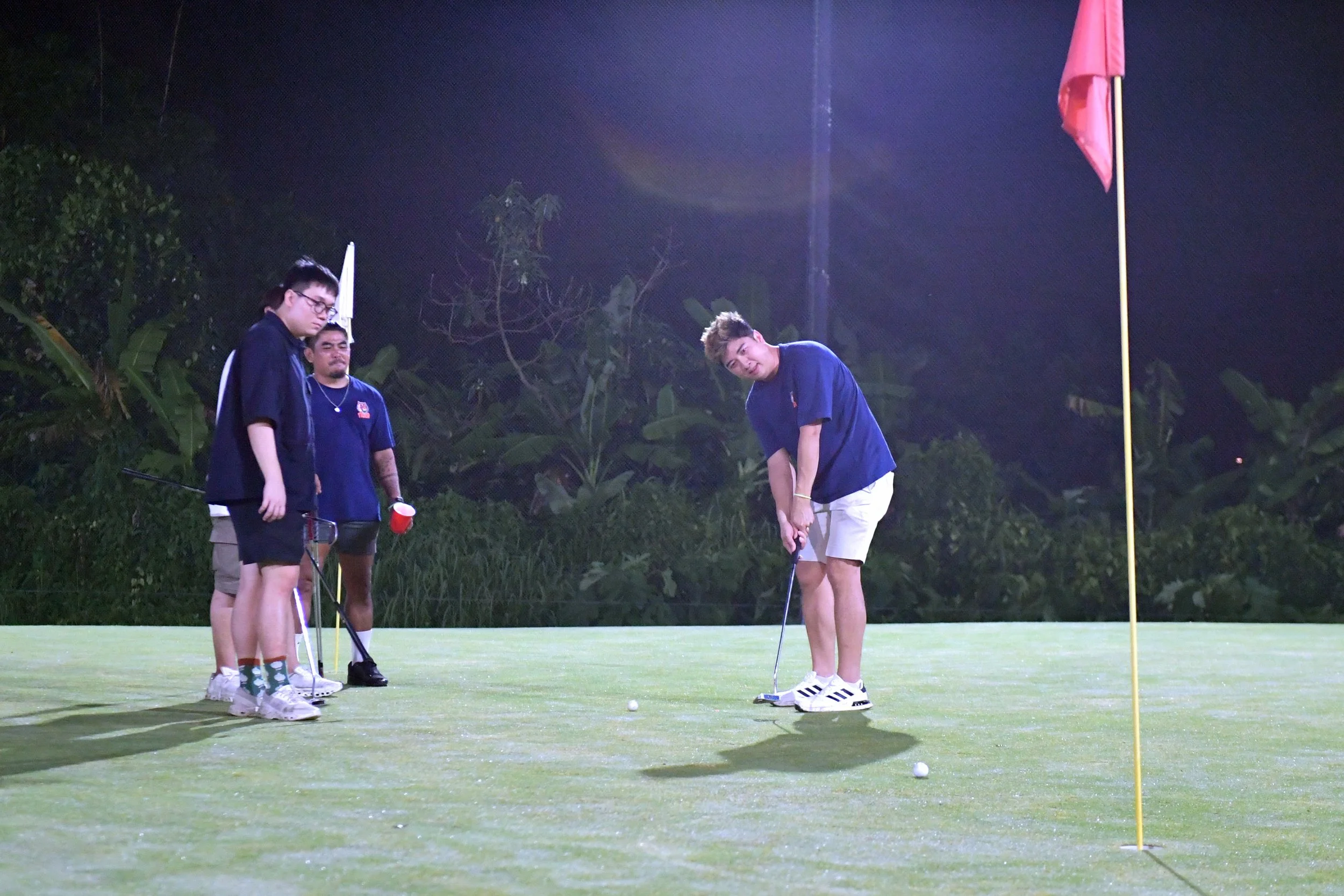 A group of young people playing golf on a night-time putting green, with one person preparing to putt and three others watching, illuminated by bright artificial lights, with a golf flag and greenery in the background.