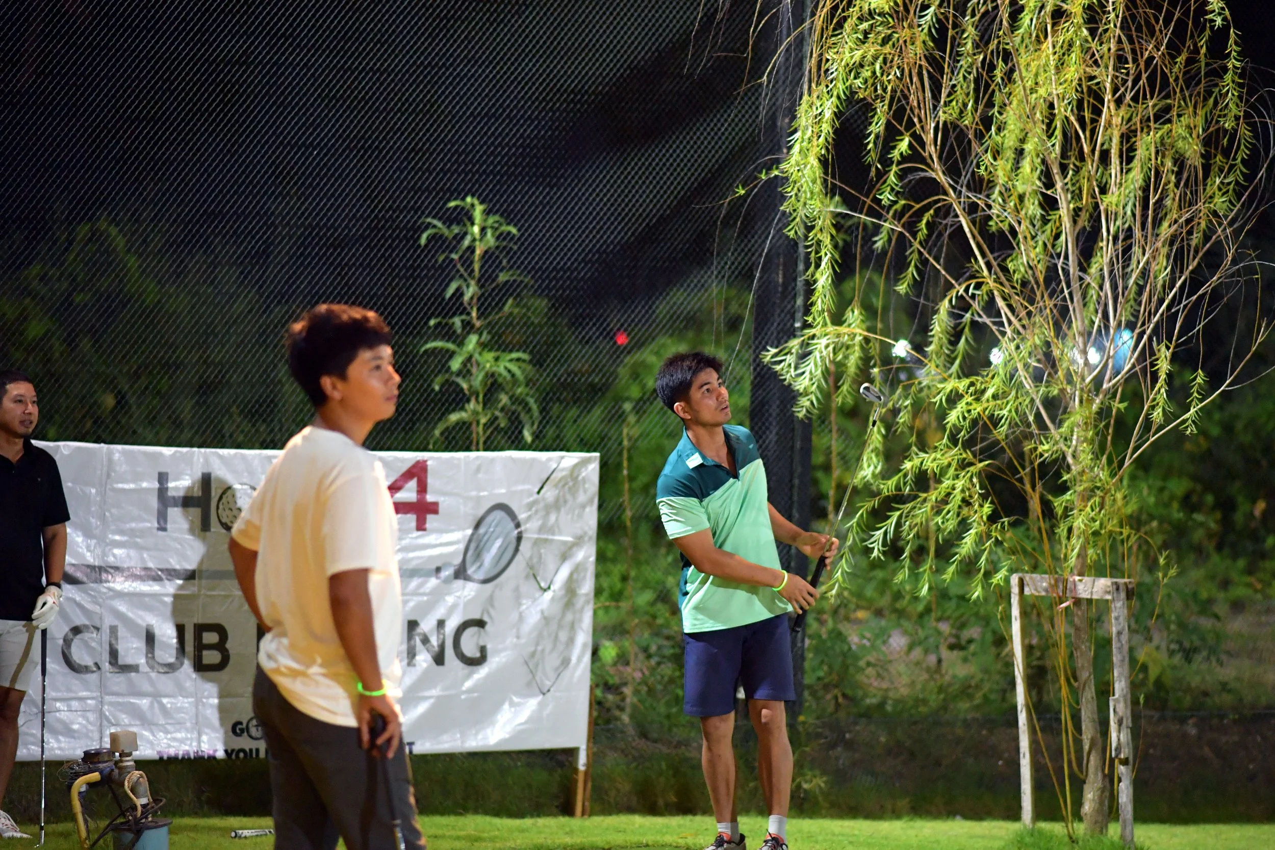 Three young men on a golf course at night, one hitting a golf ball towards a tree illuminated by outdoor lights, with a banner in the background and greenery surrounding the scene.
