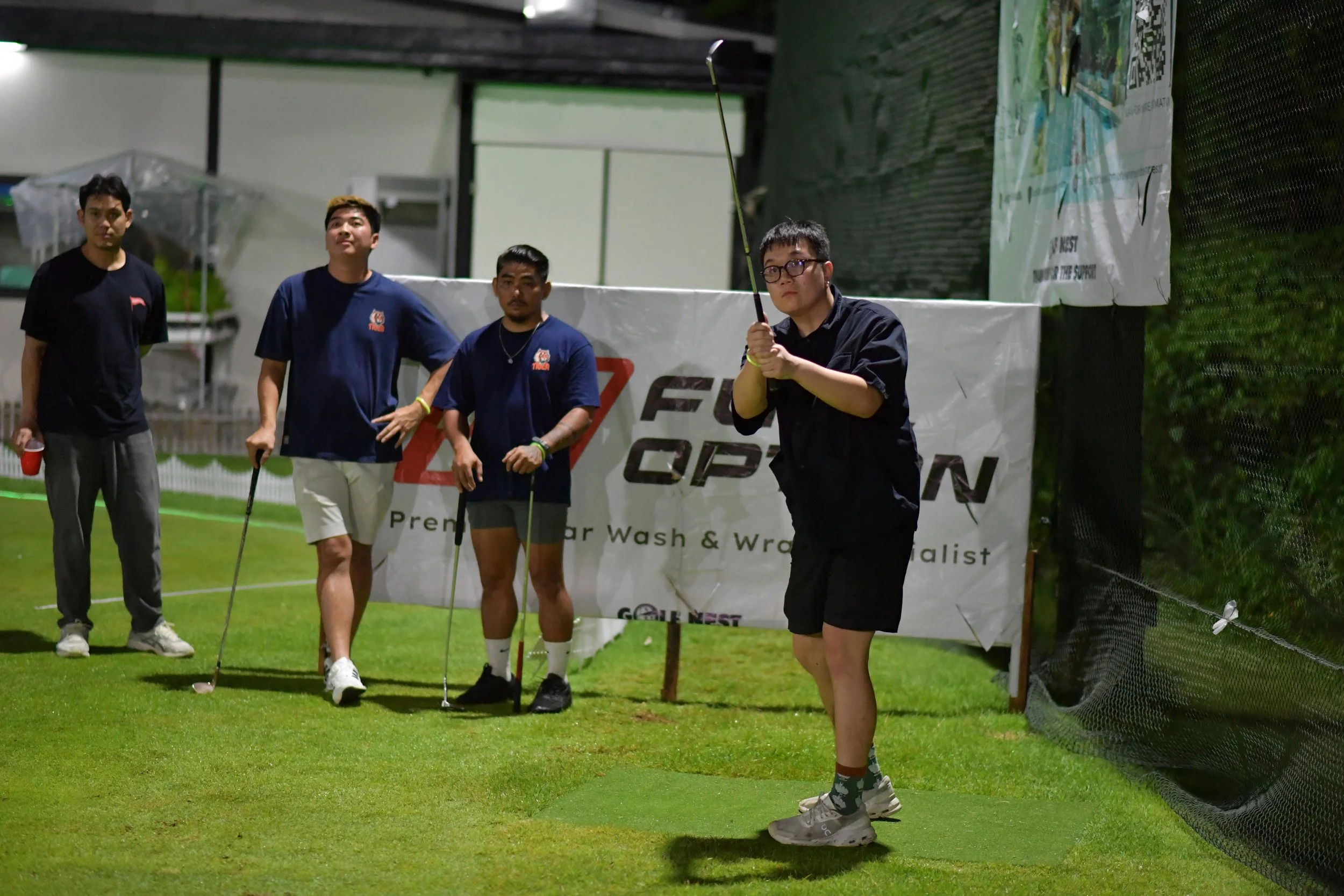 Four young men at a golf driving range, one preparing to swing a golf club, two standing with clubs, and one standing with a drink, against a backdrop of a sign and netting.