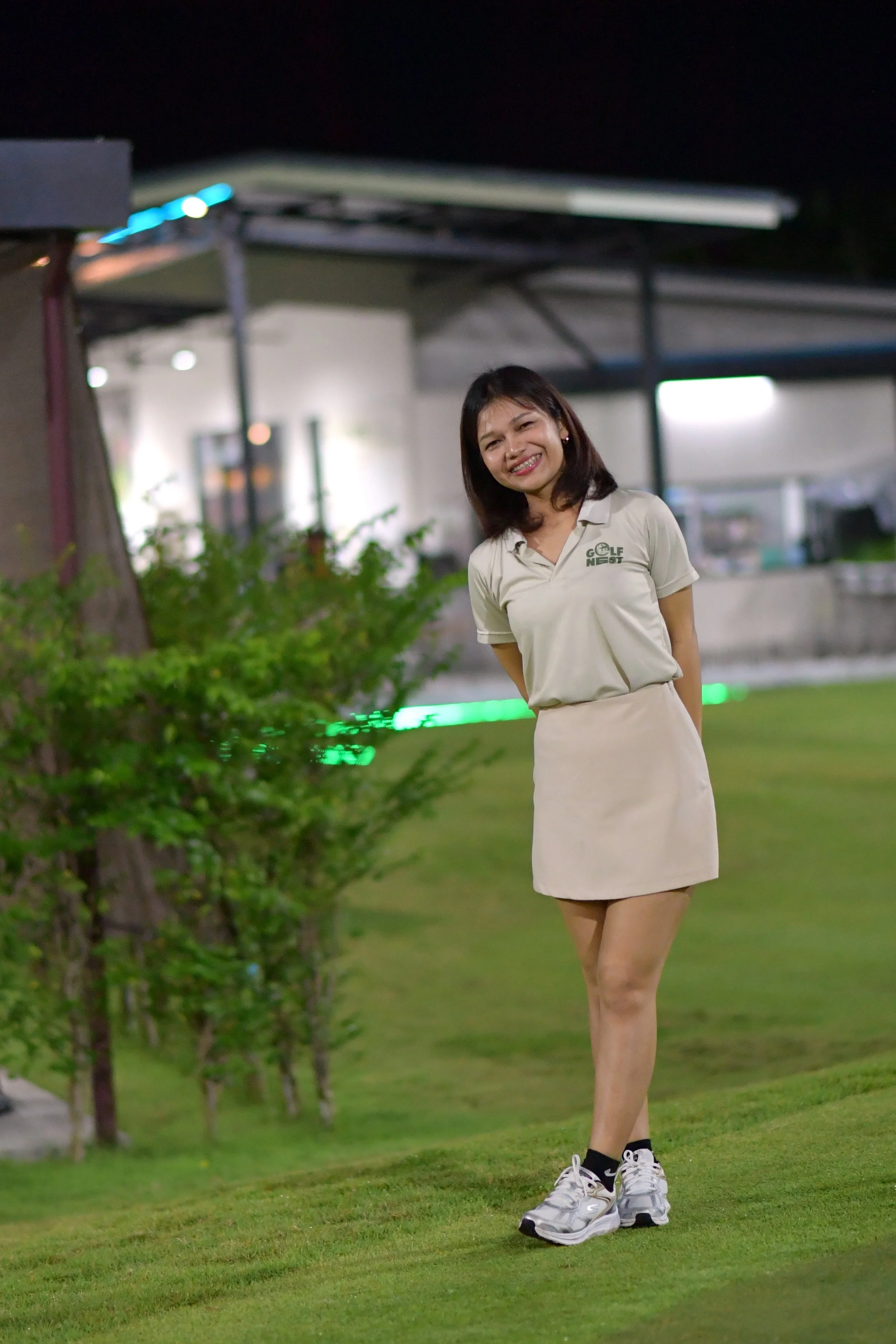 A woman in a beige golf uniform standing on the grass at night, smiling, with a building and greenery in the background.