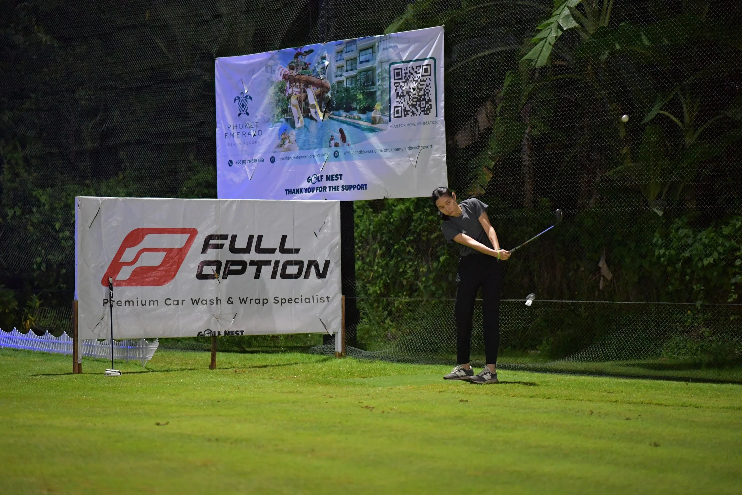 A woman playing golf on a green field, preparing to hit a golf ball, with advertising banners in the background.