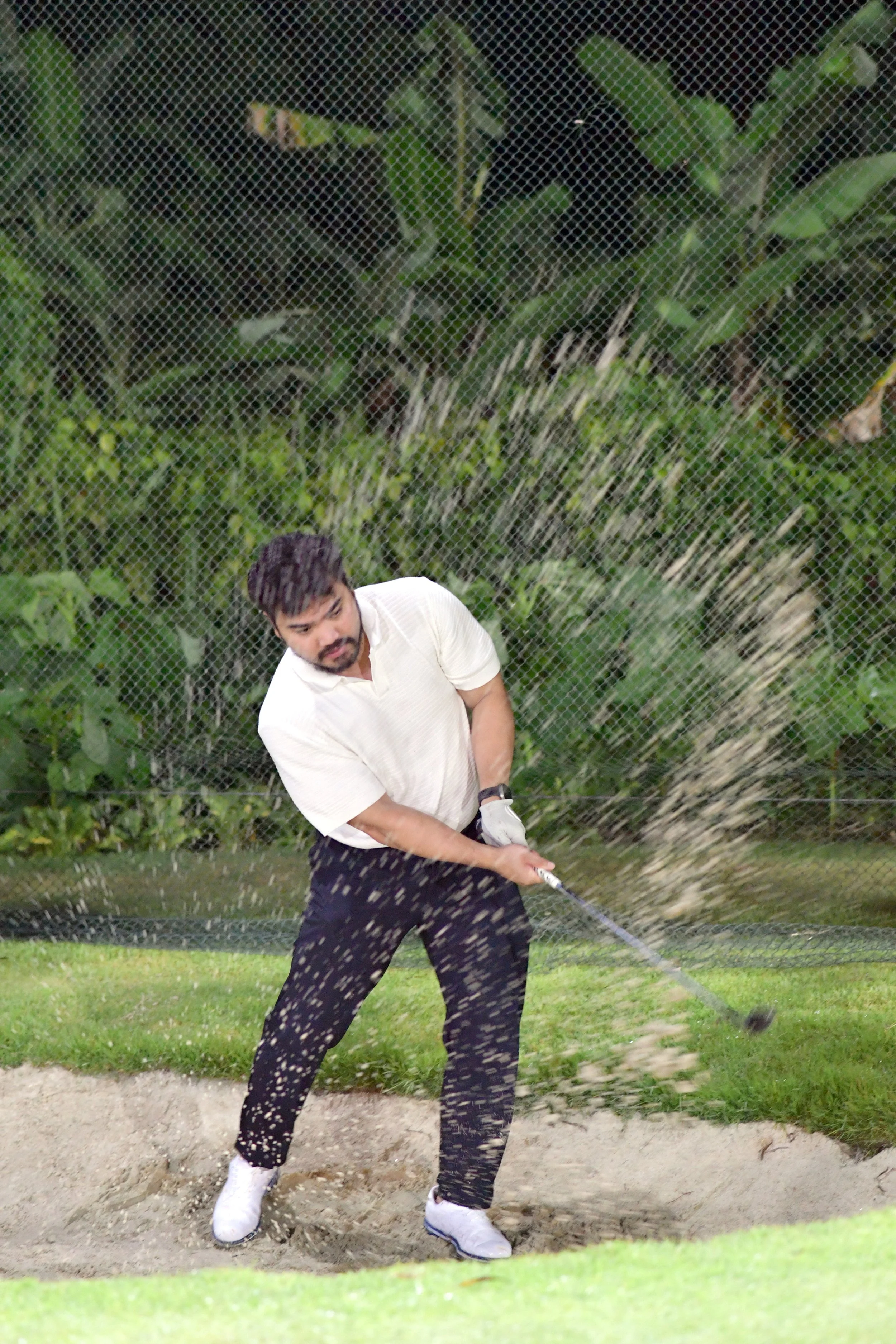A man playing golf in a sand trap, swinging a golf club with grass and trees in the background.