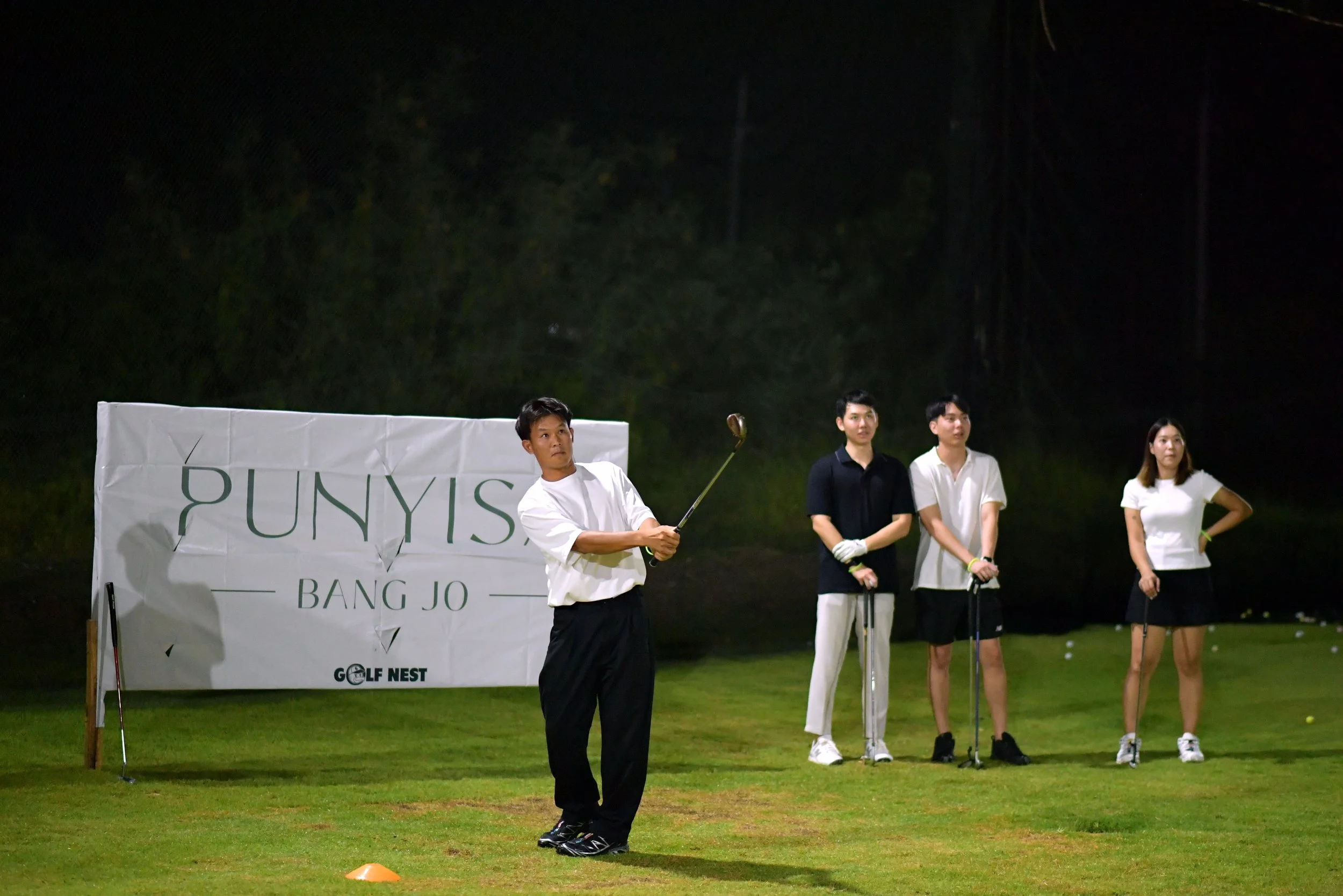 A group of people practicing golf at night on a golf course with a banner that reads 'PUNYIS BANG JO' and 'GOLF NEST' in the background.