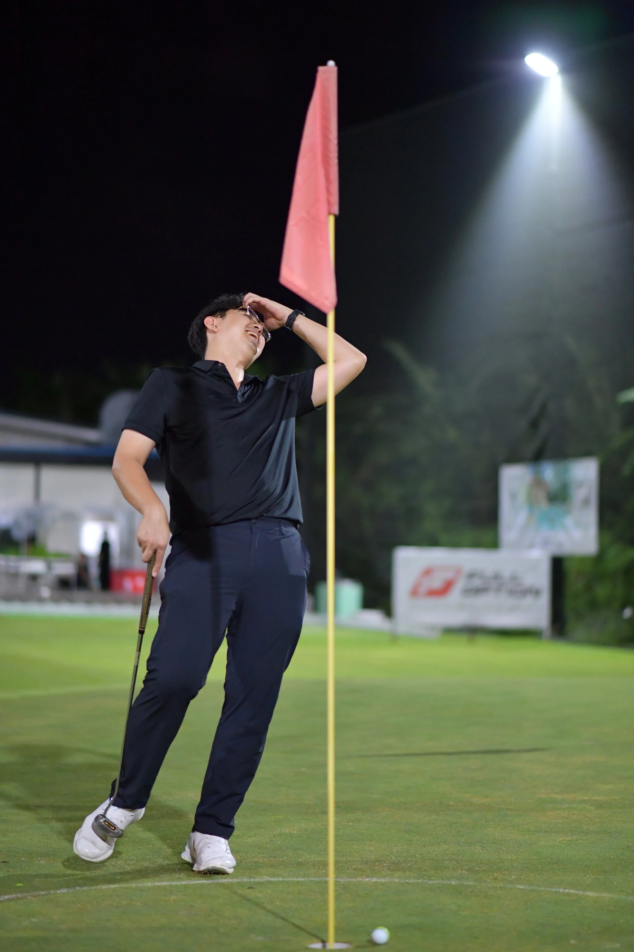 A man in a black polo shirt and navy pants on a golf course at night, holding a golf club, smiling, and standing next to a flagstick with a golf ball nearby.