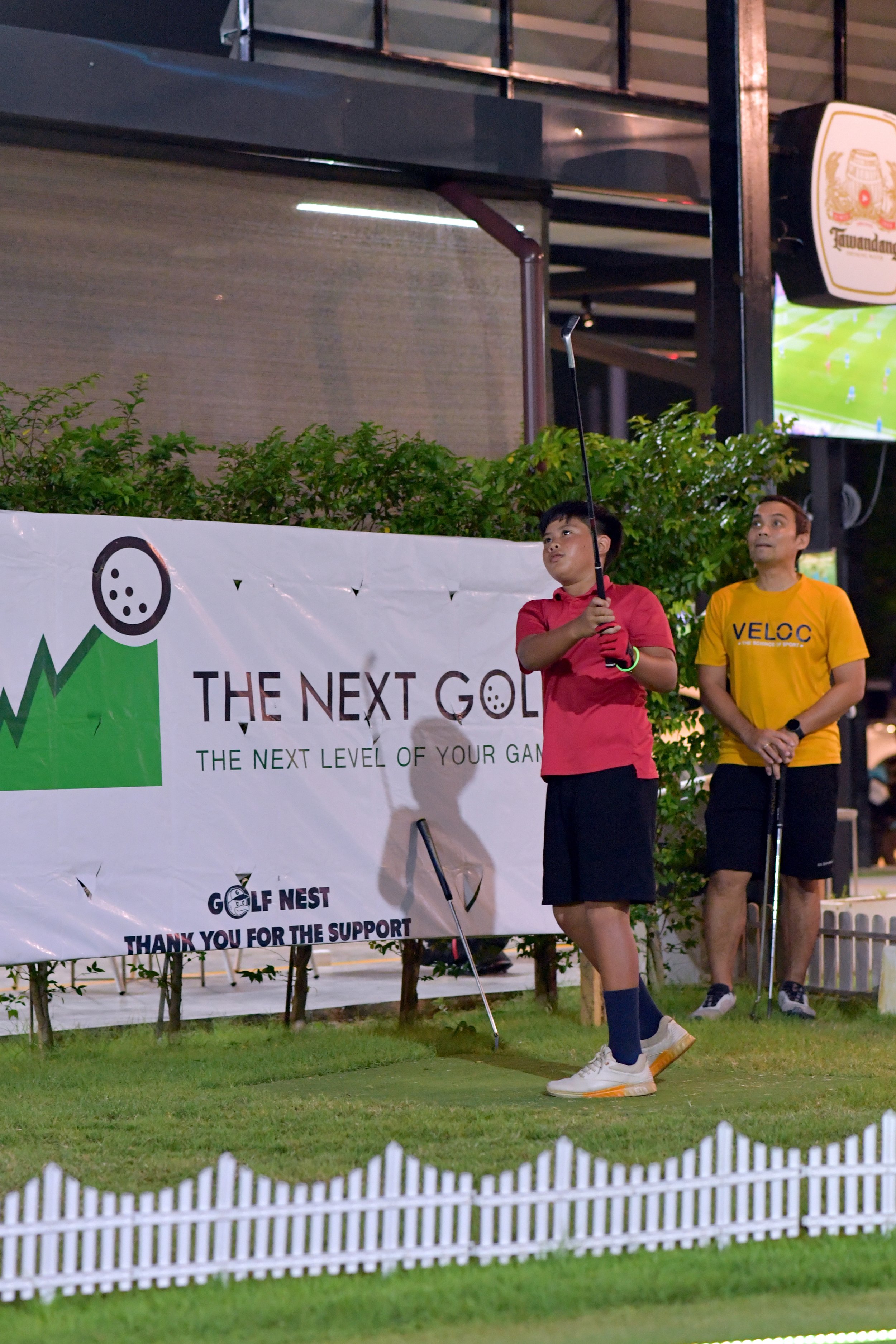 A young boy in a pink polo shirt and navy shorts is preparing to swing a golf club, standing on a small green mat at a golf practice area, with a man in a yellow shirt watching nearby, set in a well-lit outdoor setting.