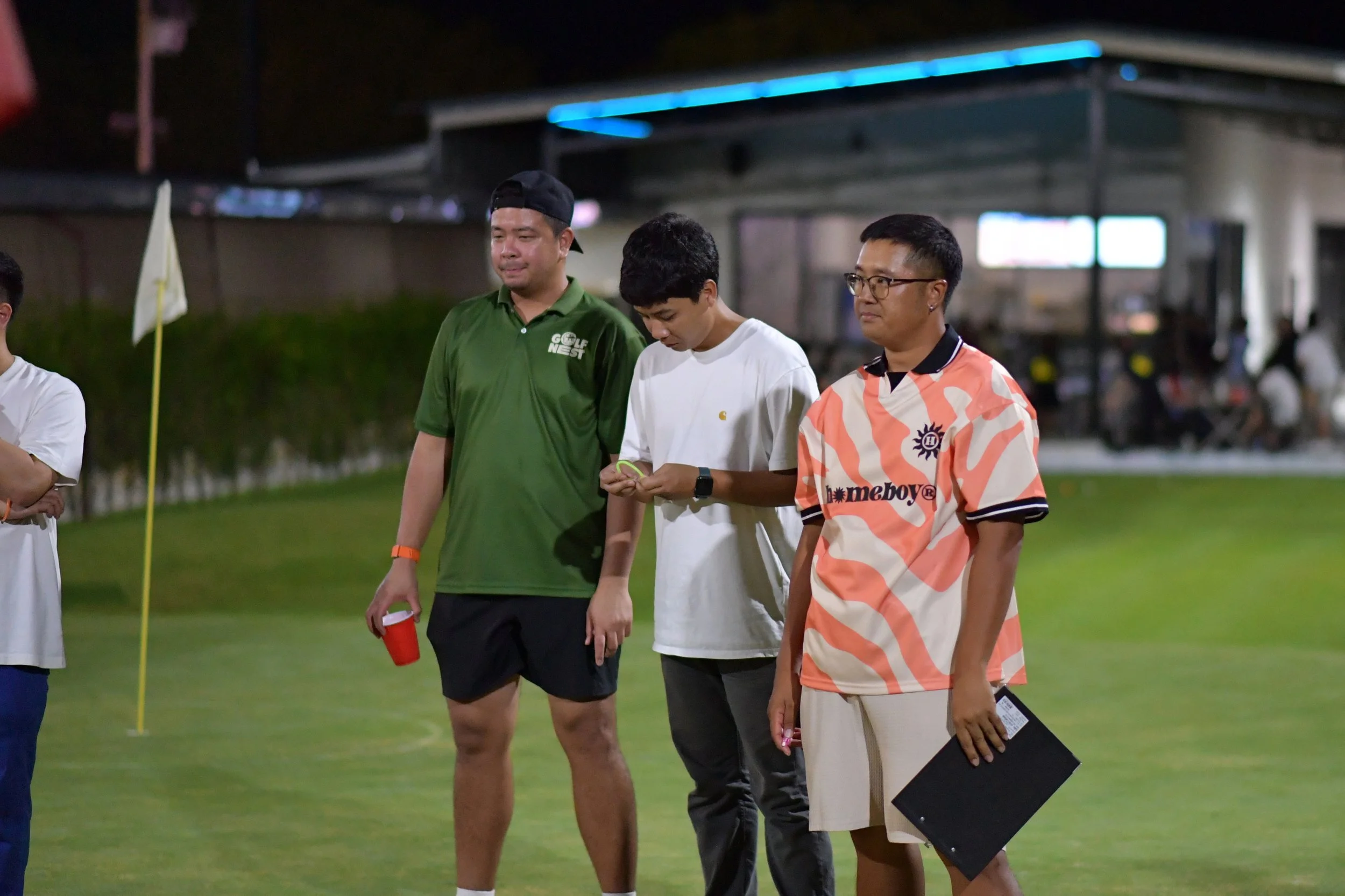 Three men standing on a golf course at night, with a flagpole and a building in the background. One man is wearing a green golf shirt and black shorts, holding a red cup. The second man is in a white t-shirt, looking down at his phone. The third man 
