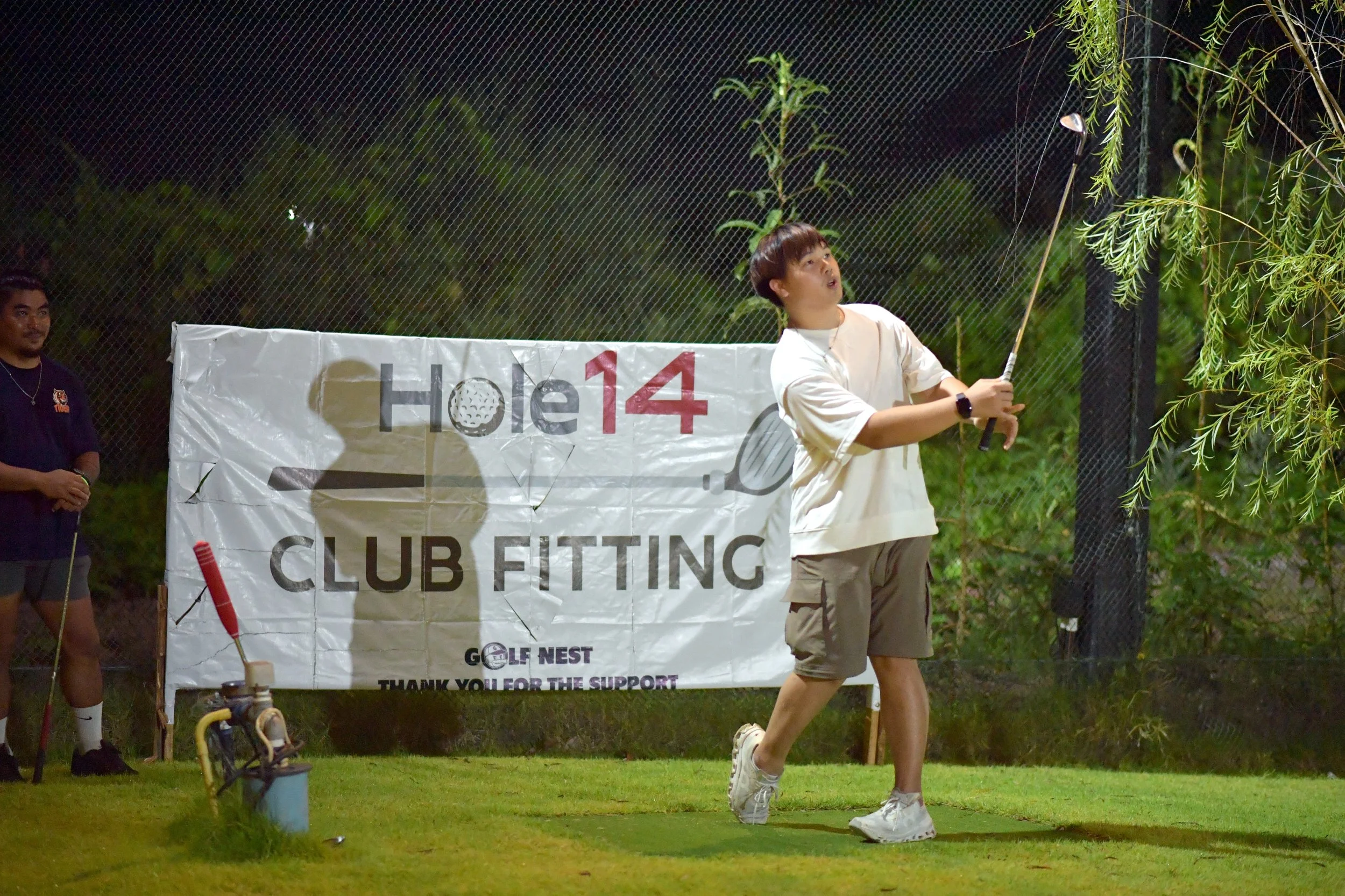 A young woman hitting a golf ball on a practice mat at night, with a golf club in her hands and a banner behind her that reads 'Hole 14 CLUB FITTING'.
