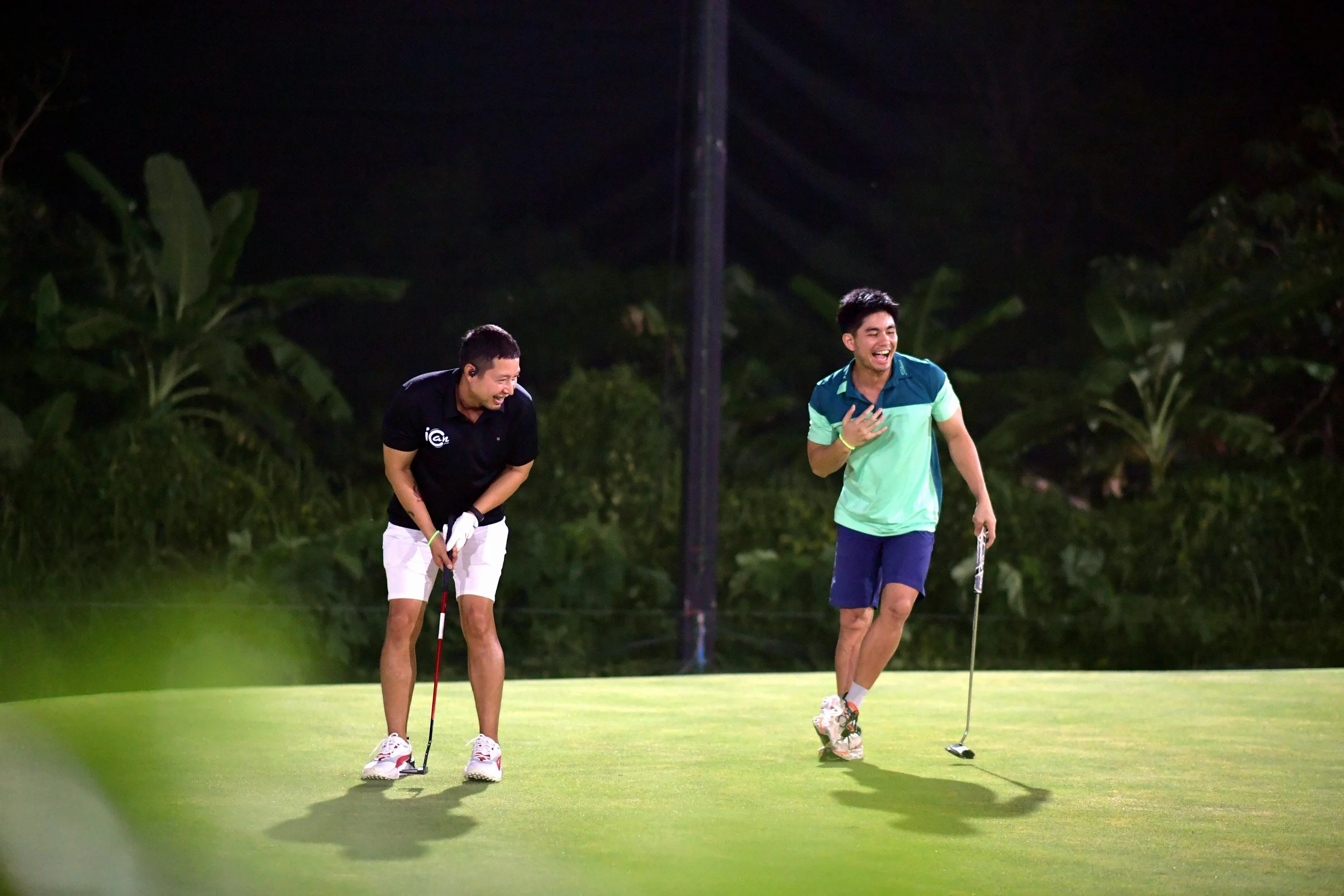 Two men playing golf on the green at night, both laughing and enjoying the moment.