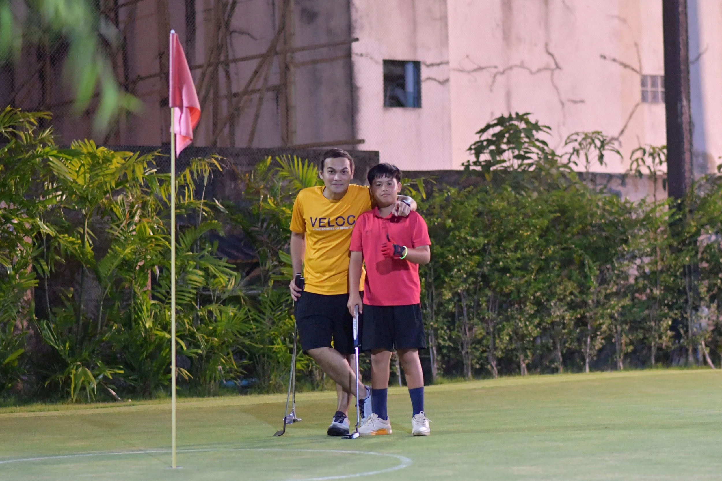 A man and a boy standing on a golf course near a flag, with grassy areas and trees in the background.