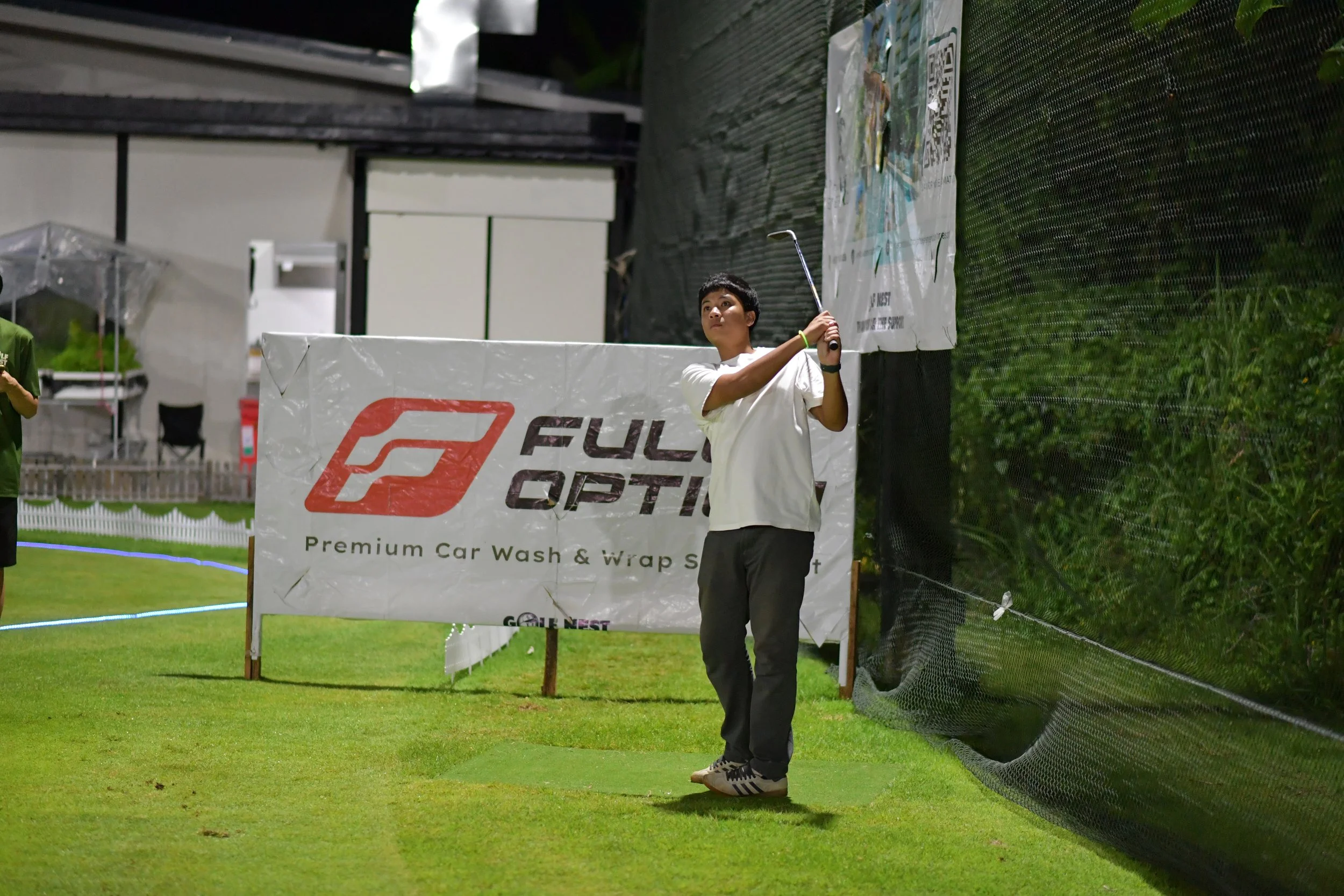 A young man in a white shirt and dark pants practicing golf swing at an indoor golf practice range at night. He is swinging a golf club near a black net and green grass, with a large advertising banner in the background.