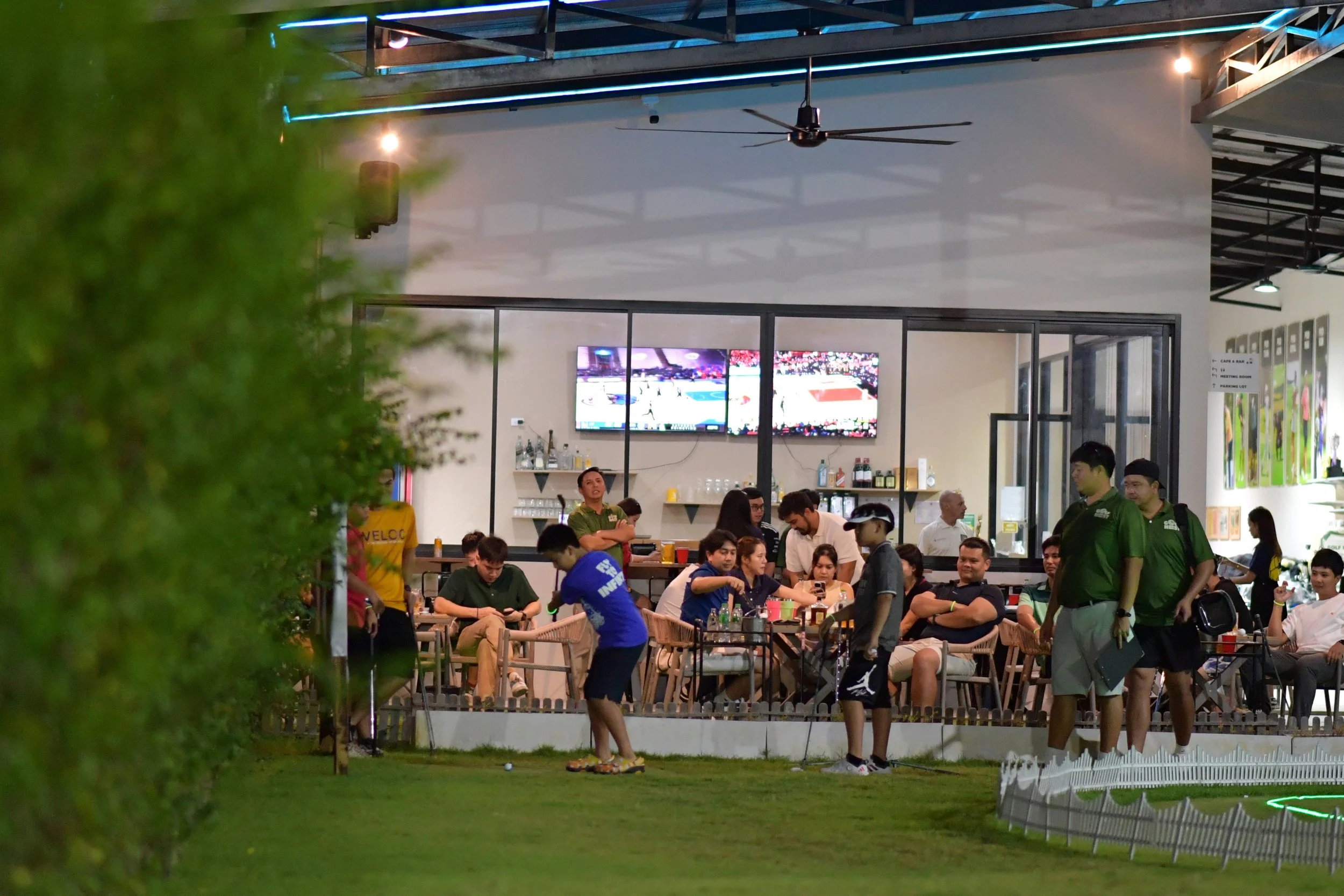 Indoor gathering at a restaurant or cafe with people seated at tables and a few individuals standing. People are playing mini-golf on the outdoor grass area in the foreground. The interior has a bar with bottles and glasses, large screens displaying 