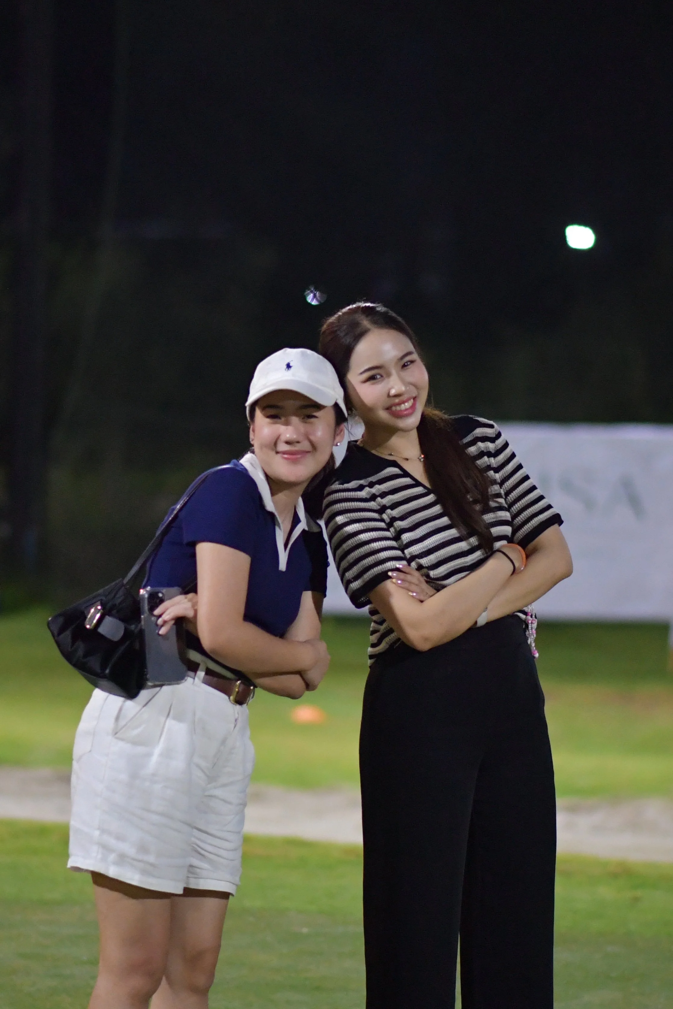 Two women standing close together outdoors at night, smiling at the camera, with one wearing a white cap and polo shirt, and the other in a striped shirt and black pants.