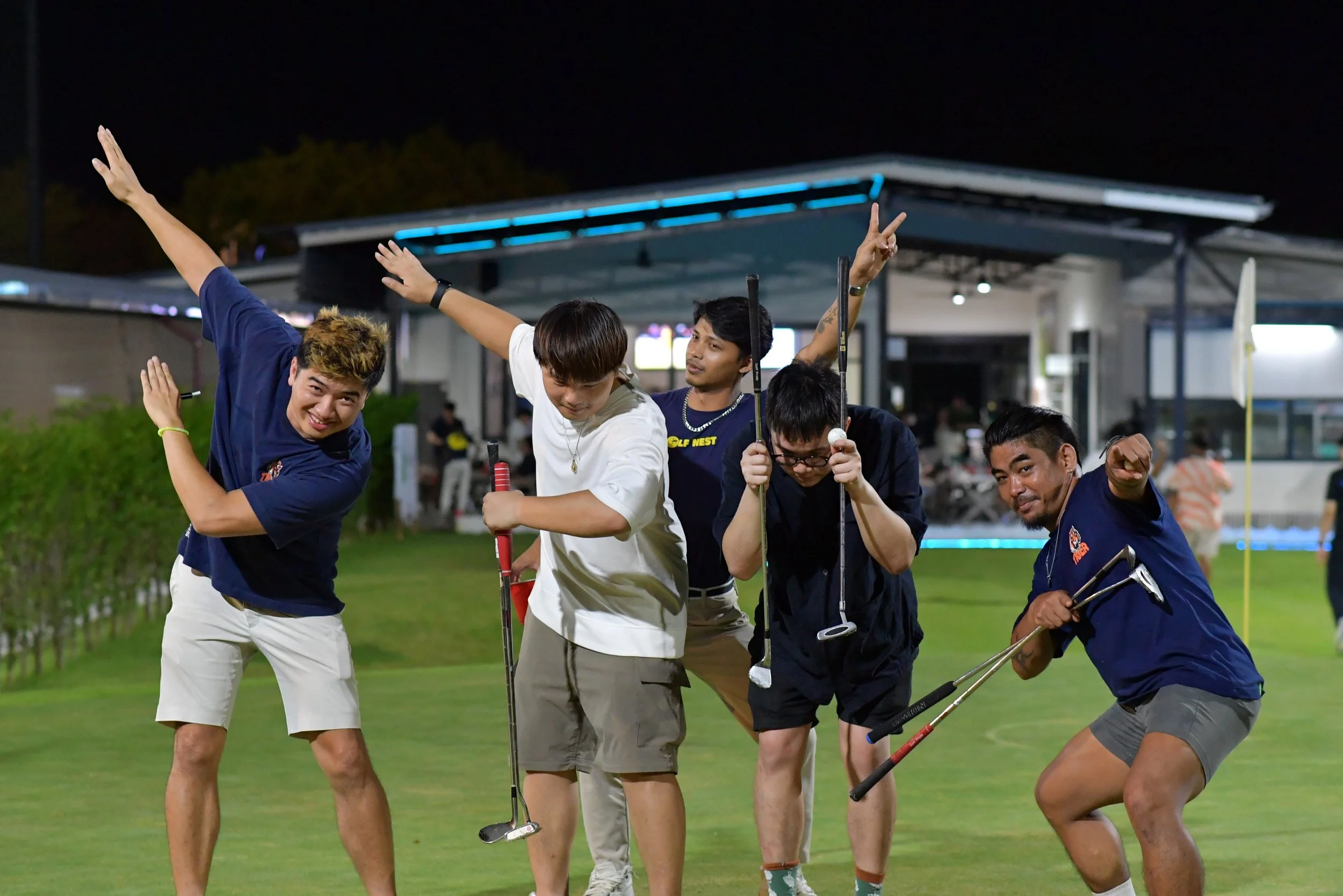 Five young men on a golf course at night, posing and smiling for the camera, with some holding golf clubs and making playful gestures.