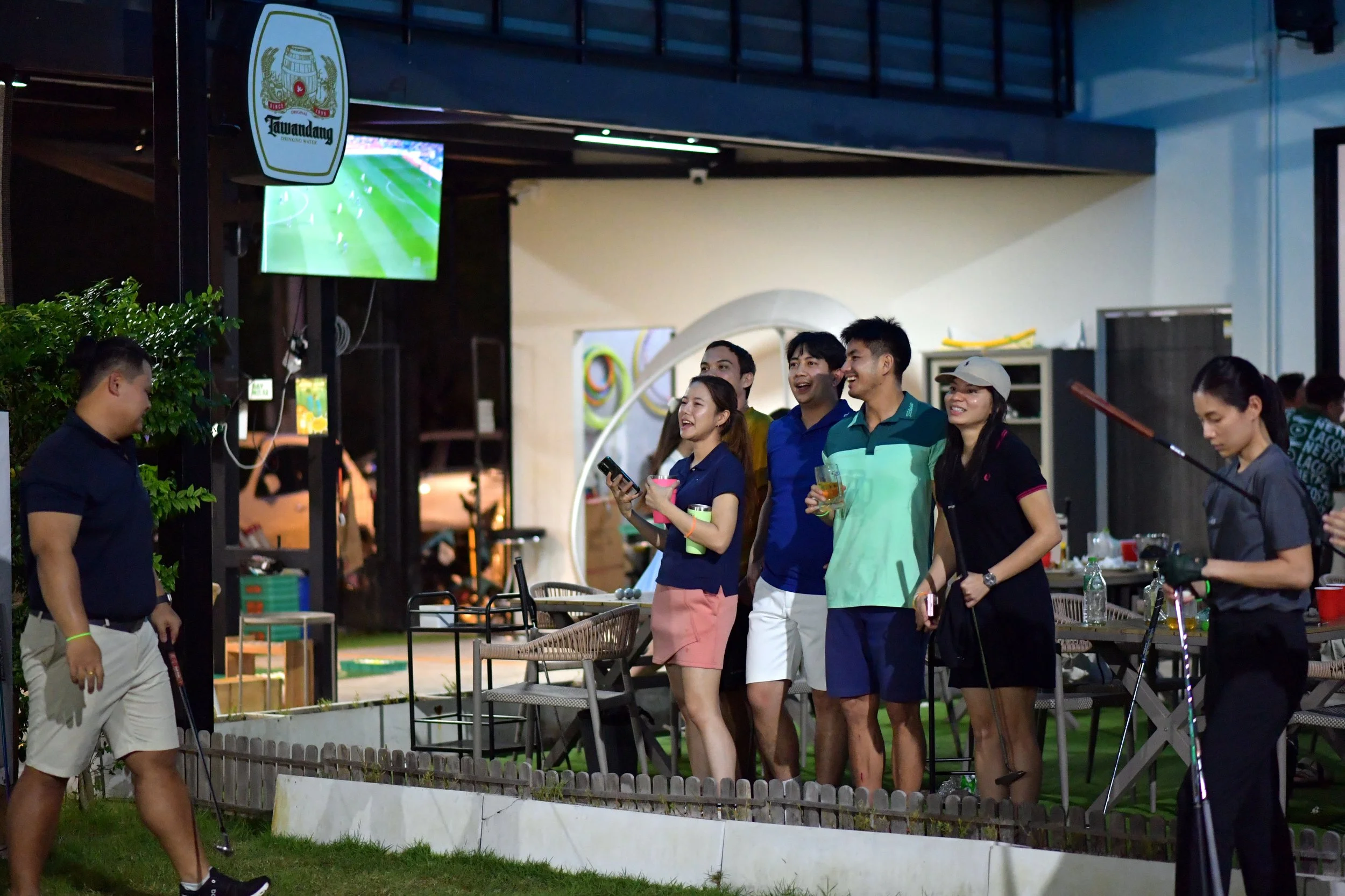 A group of friends watching a golf game on TV at an outdoor bar or restaurant during the evening, with one person preparing to take a shot with a golf club.