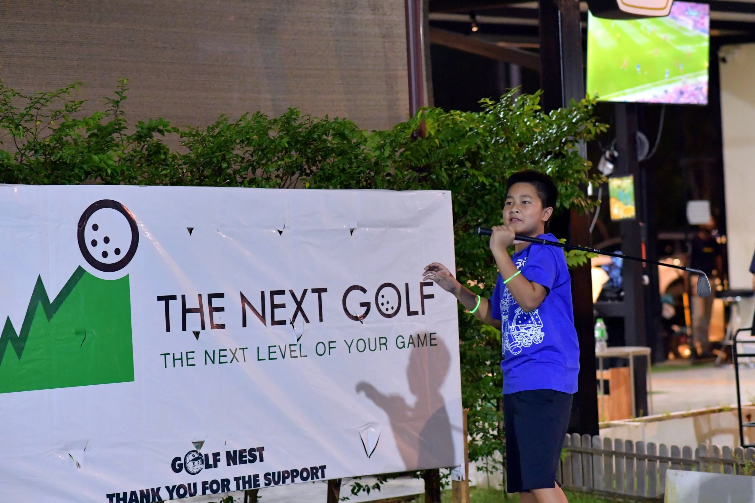 A young boy in a blue shirt holding a golf club stands next to a banner that reads 'The Next Golf: The Next Level of Your Game.' The scene appears to be outdoors at night, with a TV screen visible in the background.