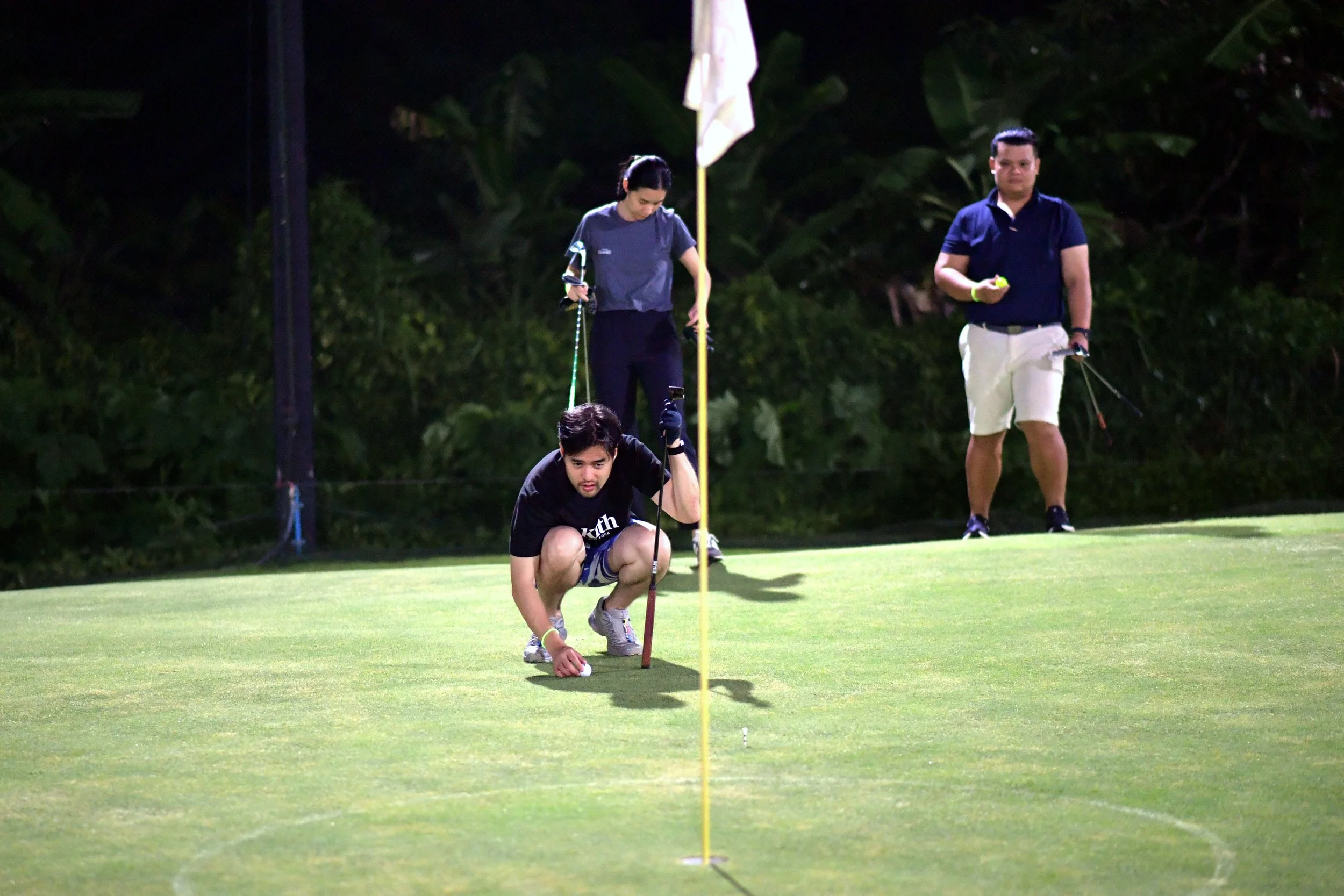 Group of people playing golf at night on a green field with trees in the background.