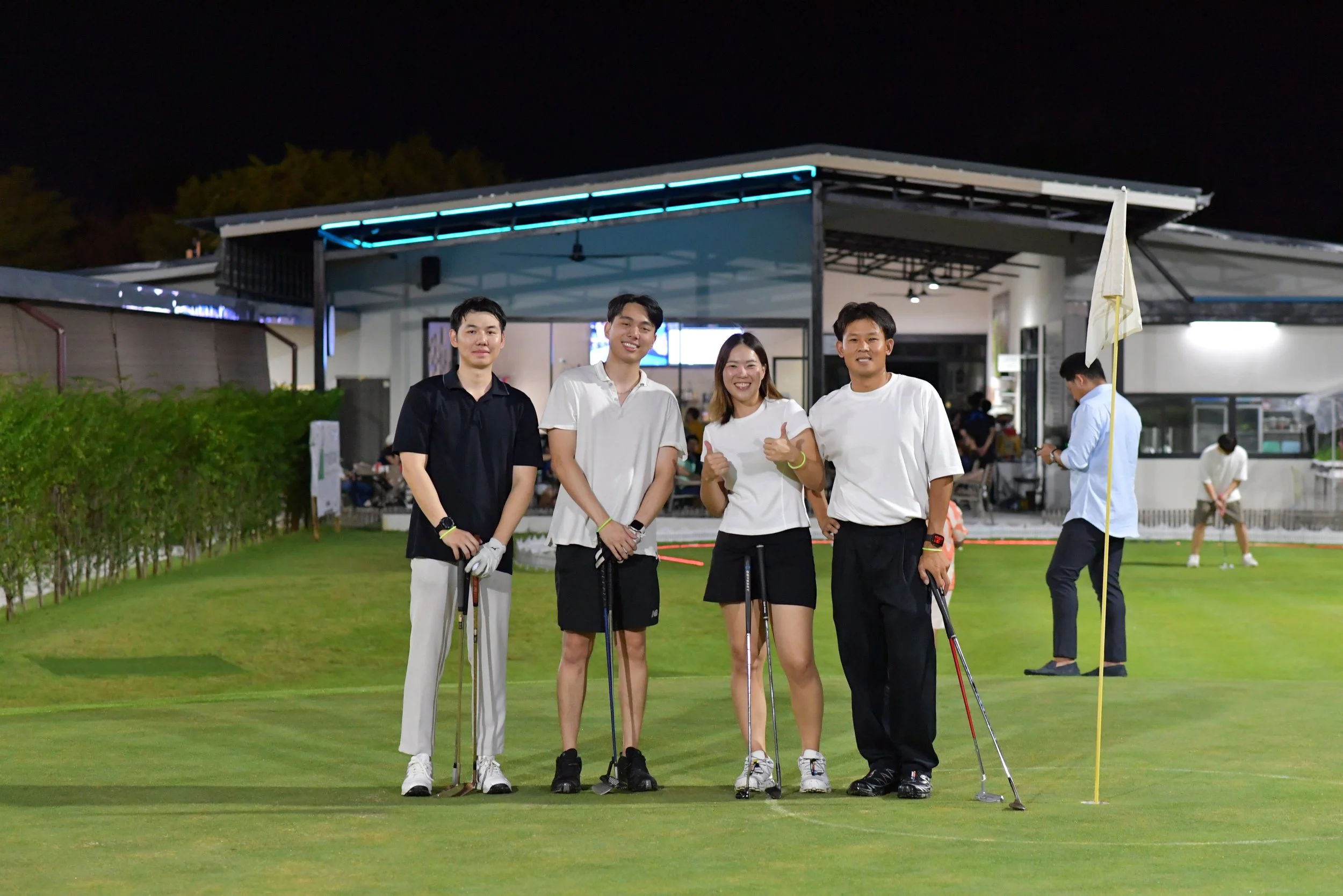 Group of four young adults posing with golf clubs on a golf course at night, smiling and making thumbs-up gestures, with a small building and another person in the background.