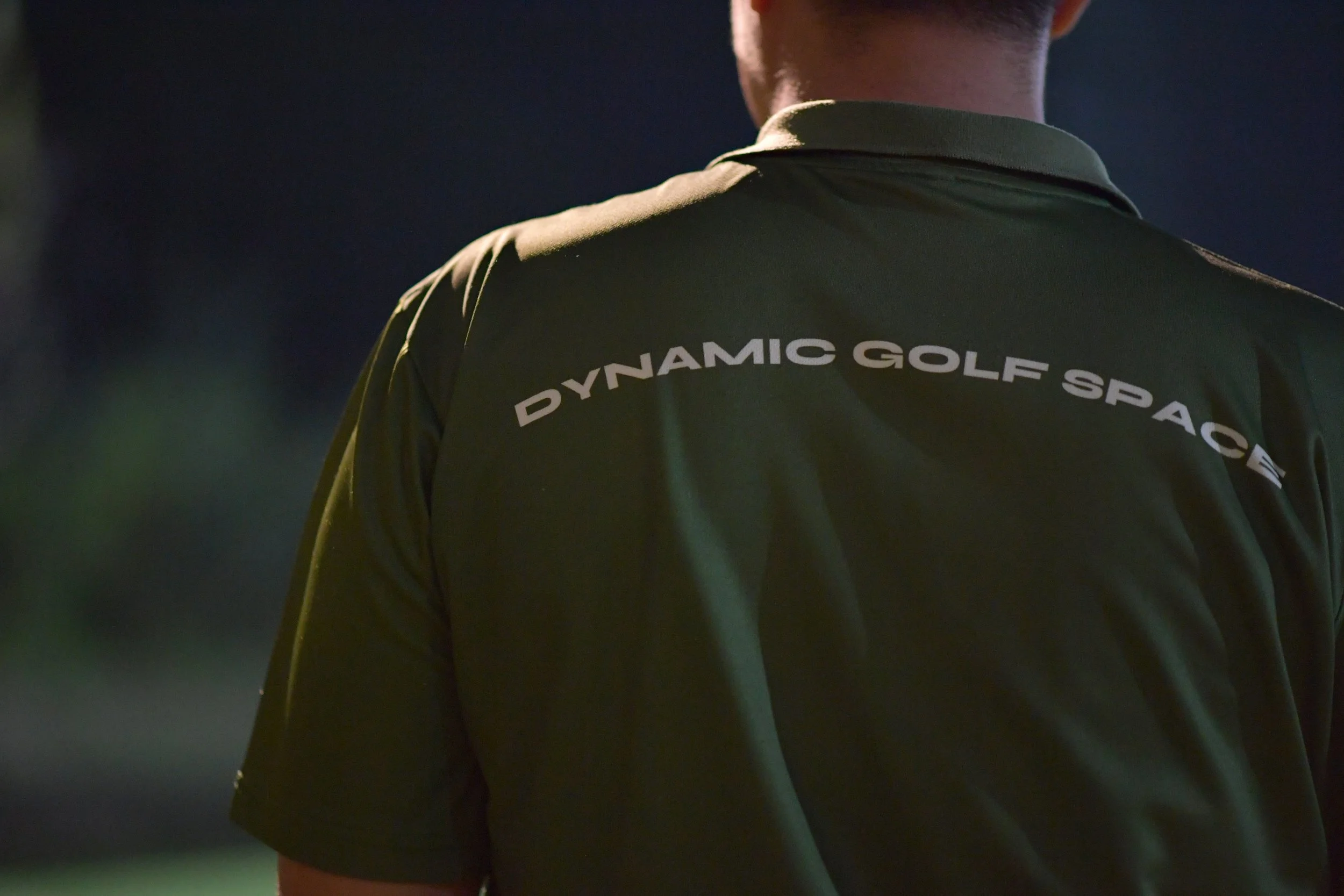 Back of a person wearing an olive green polo shirt with 'DYNAMIC GOLF SPACE' written on the back, standing outdoors at night.