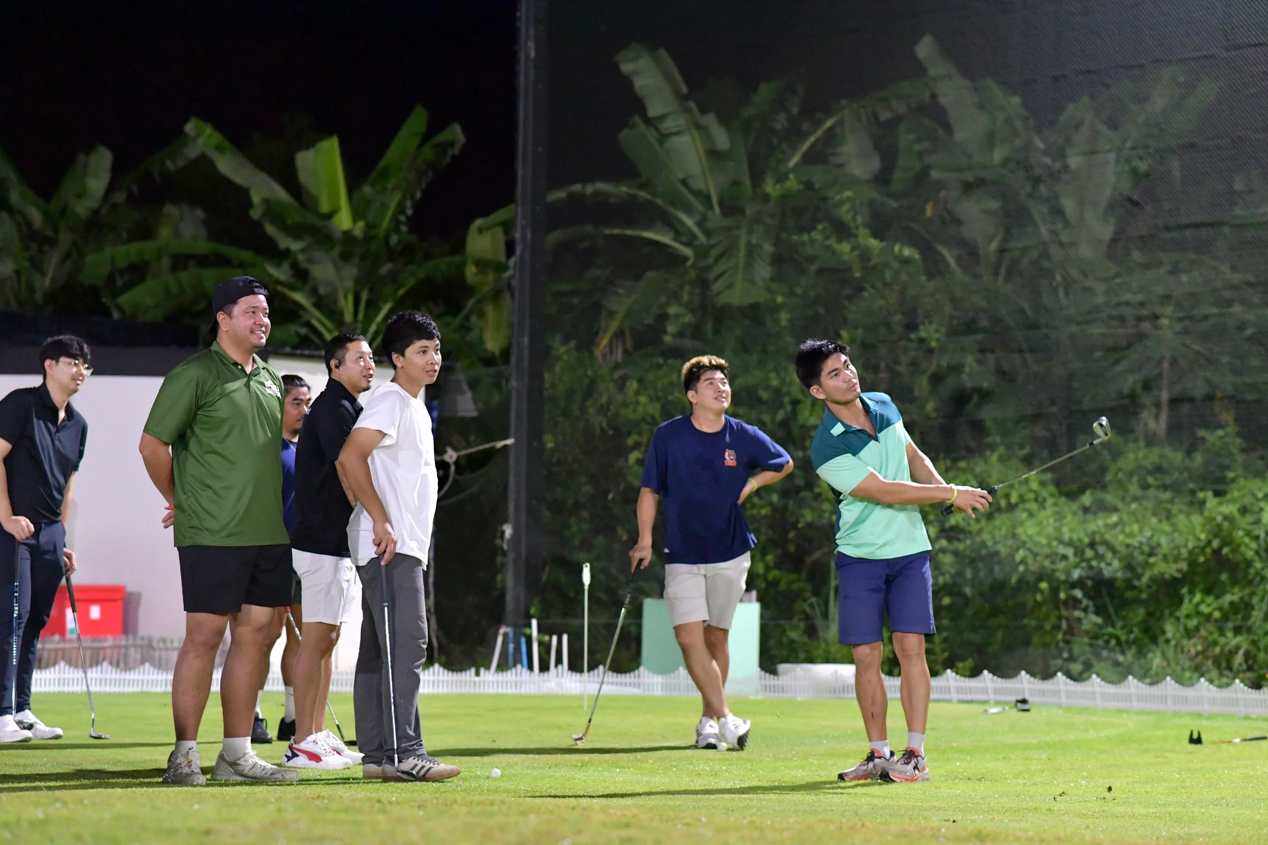 A group of seven men playing golf on a putting green at night, with one man actively swinging a golf club while others watch.