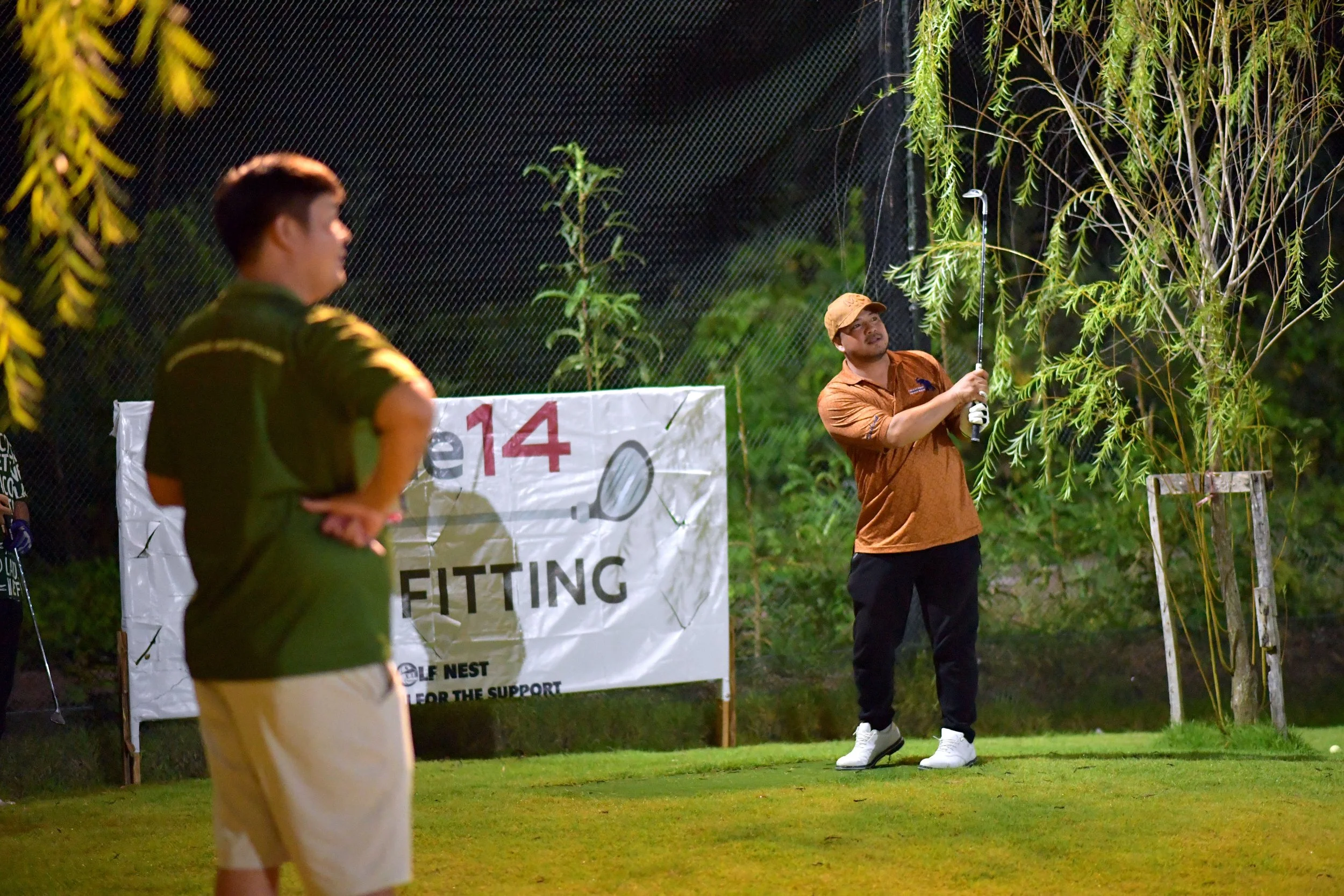 A group of people playing golf at night, one person swinging a golf club while others watch, with a sign that reads 'FITTING' and surrounded by green trees.