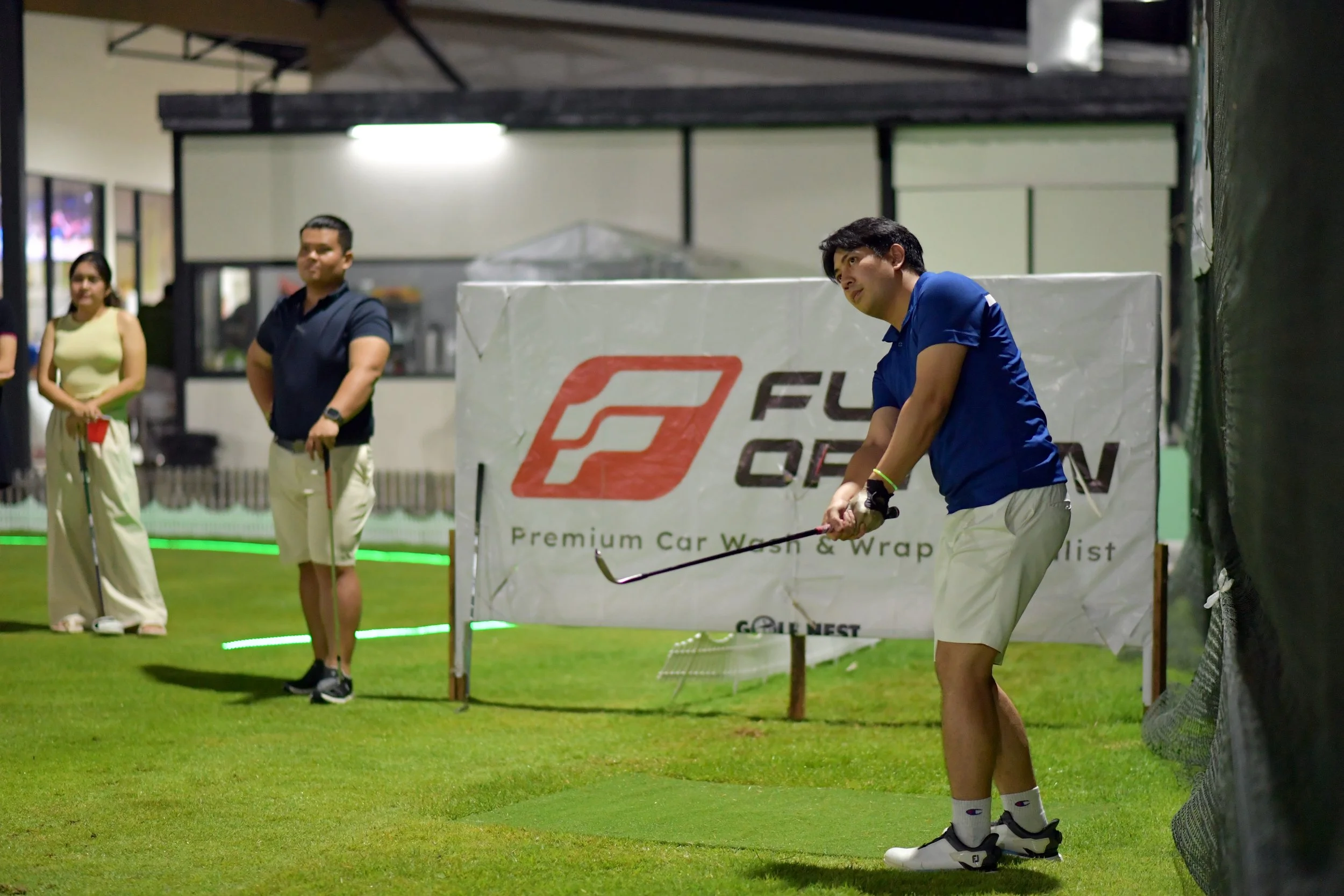 A man in a blue shirt and white shorts practicing golf indoors, with three onlookers watching him, on a grassy mat near a banner that reads 'Full Ocean.'