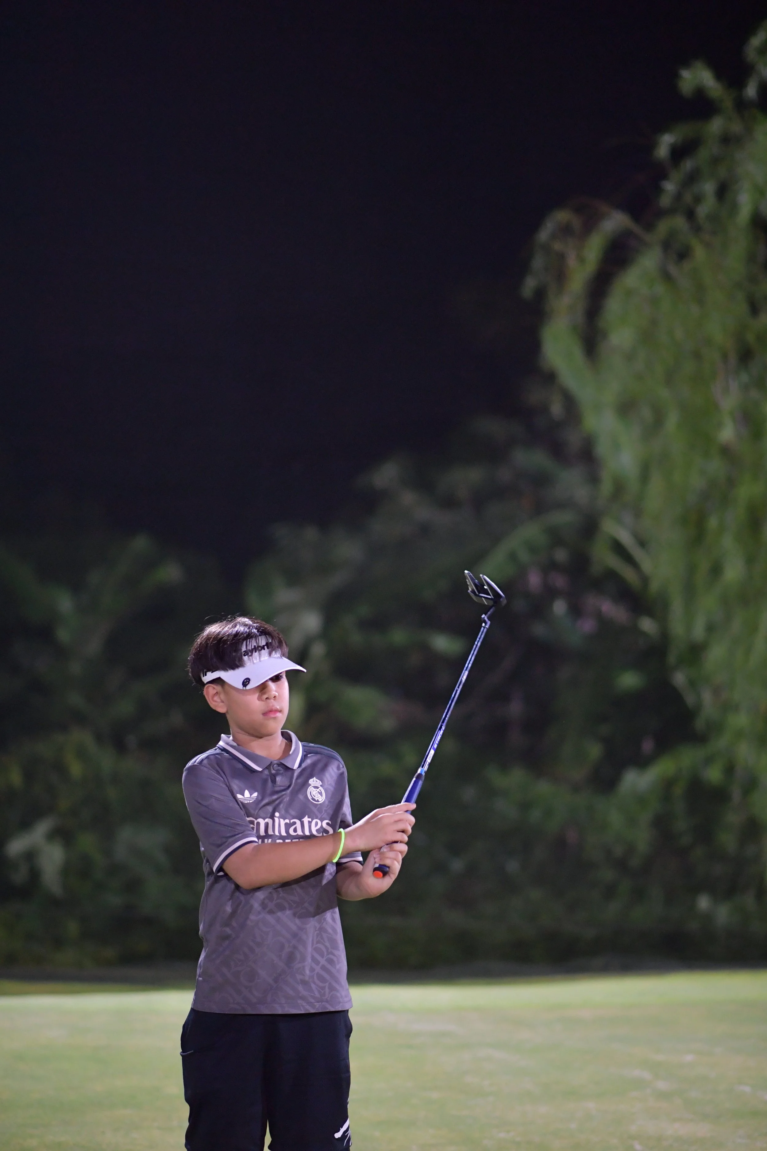 A young boy at night on a golf course, wearing a visor and a gray shirt with the Real Madrid logo, holding a golf club.