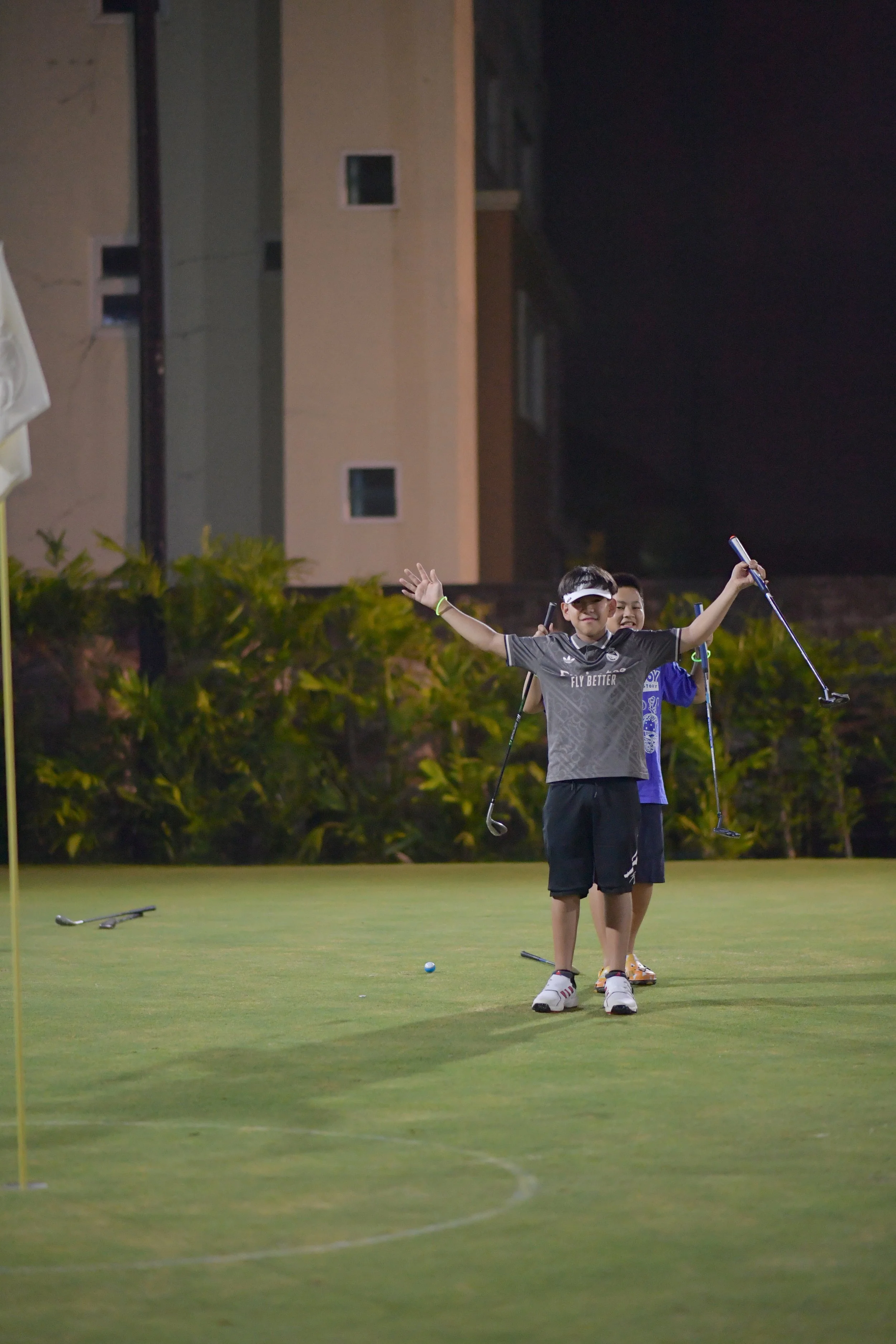Two boys playing golf on a putting green at night, with one excitedly raising his arms and holding a golf club, while the other stands behind smiling.