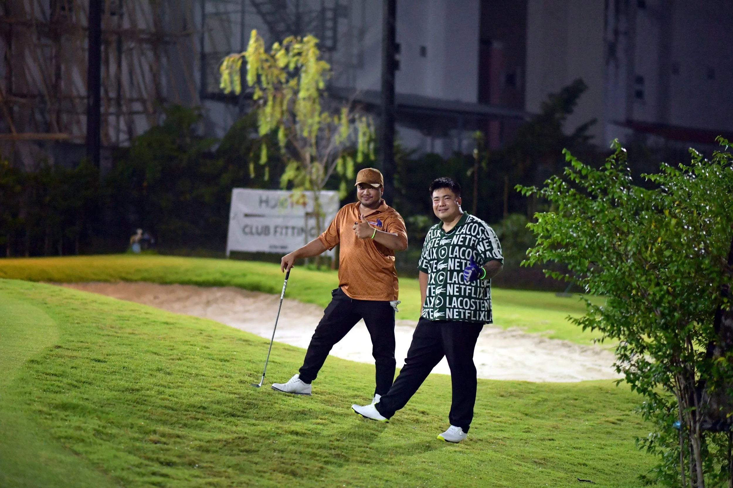 Two men standing on a golf course at night, one holding a golf club and giving a thumbs-up, the other smiling and holding a golf ball, with a sand trap and greenery in the background.