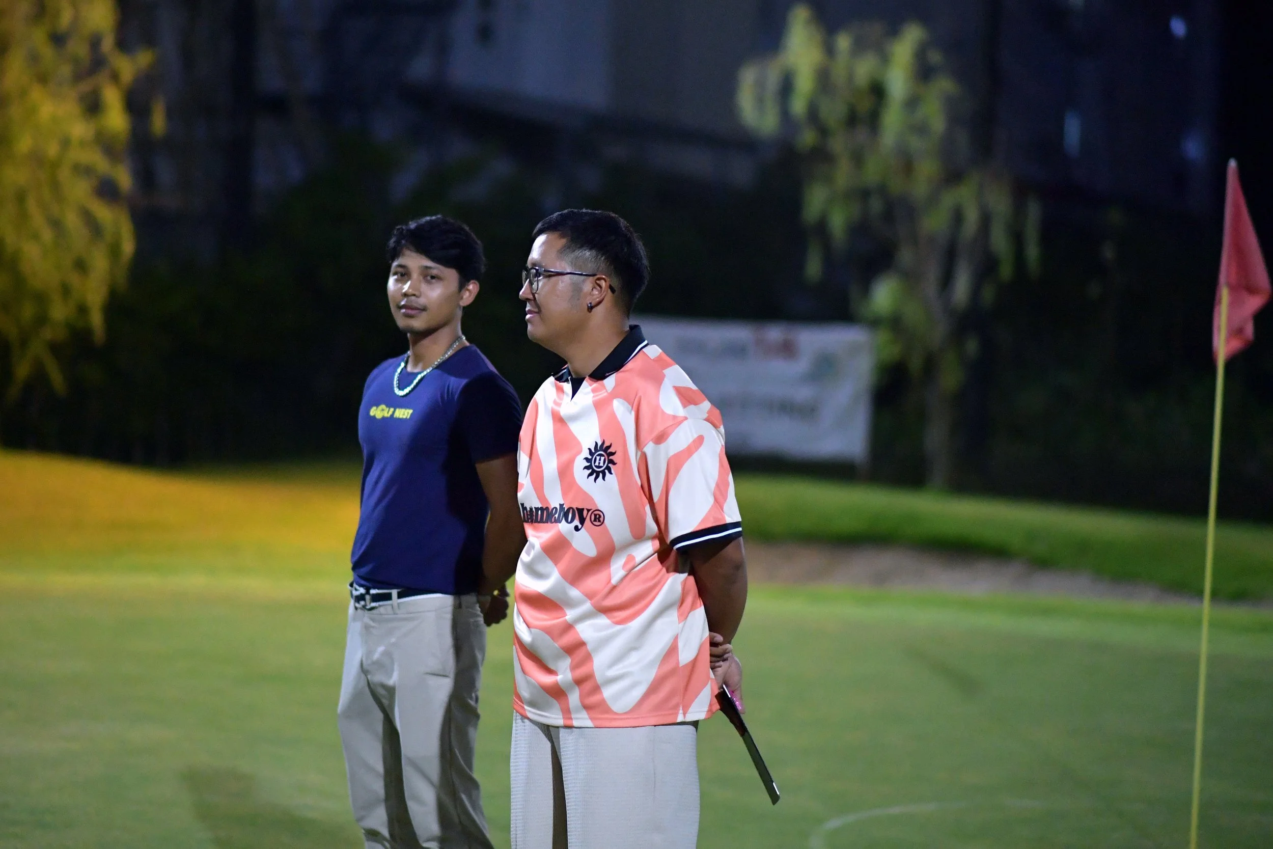 Two young men standing on a golf course at dusk. One is wearing a navy blue t-shirt and beige pants, the other is wearing a patterned golf shirt and light-colored pants, holding a club. There is a red flag on the right.