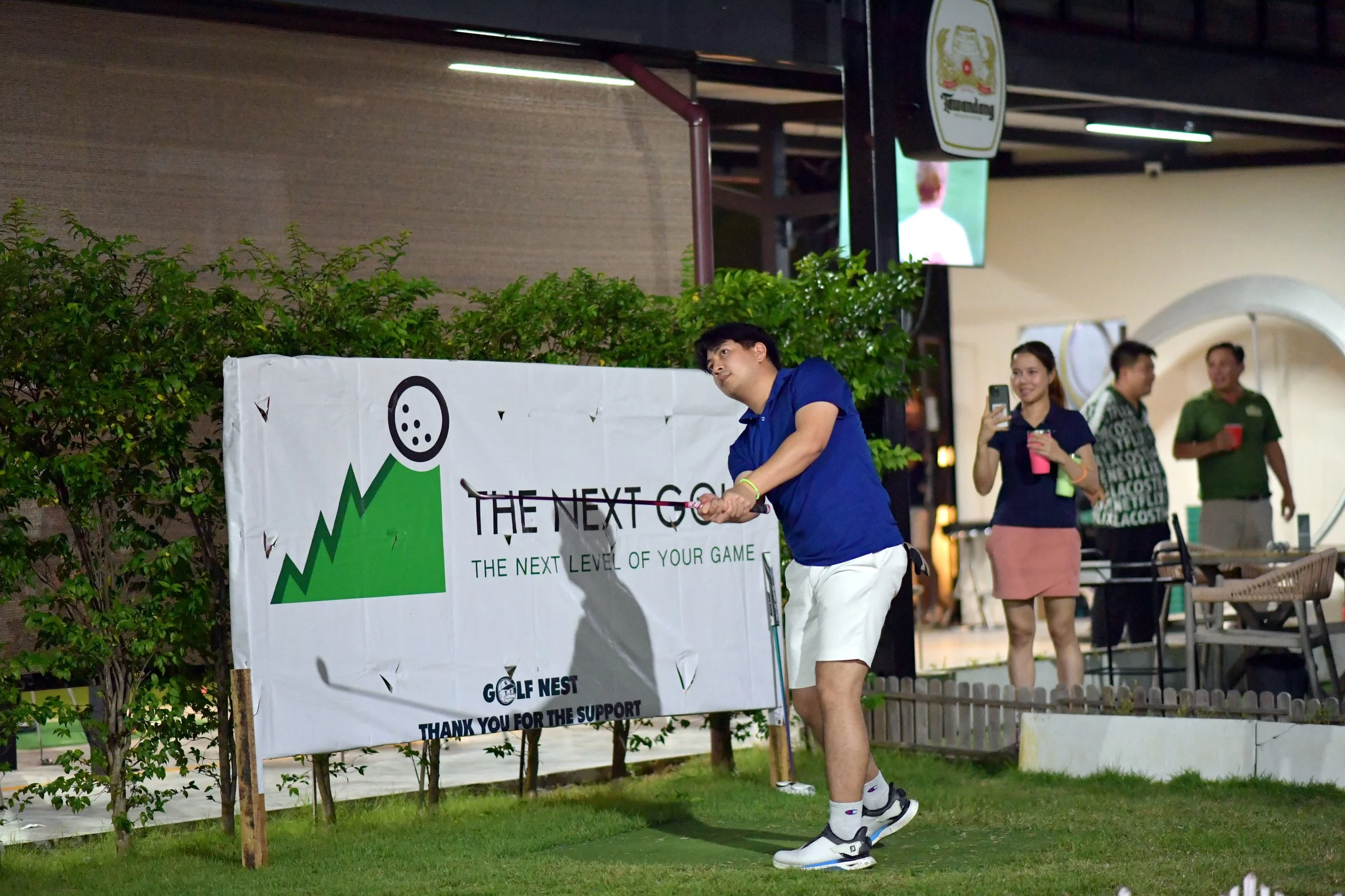 A man in a blue shirt and white shorts playing mini golf at an outdoor golf course during the night, with other people in the background taking photos and socializing.