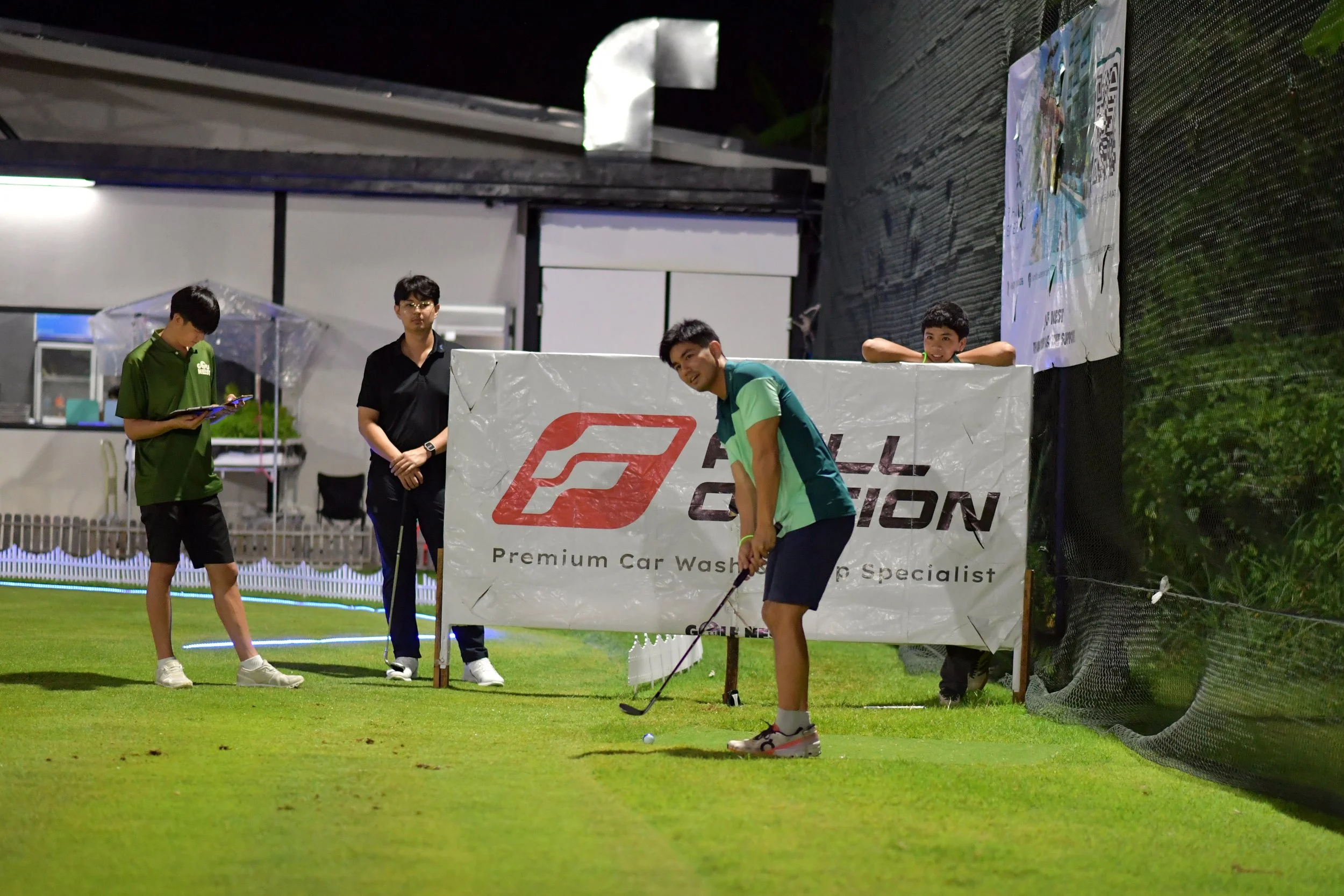 Two boys playing golf at night on a grassy field, with one preparing to hit the ball and the other observing near a crowd of three young men standing behind a banner promoting a car wash business.