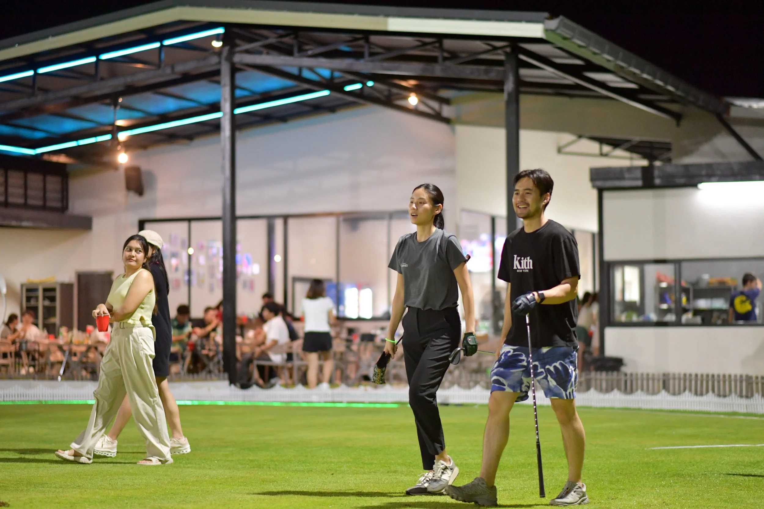 People playing golf on a green field at night, with indoor dining visible in the background.