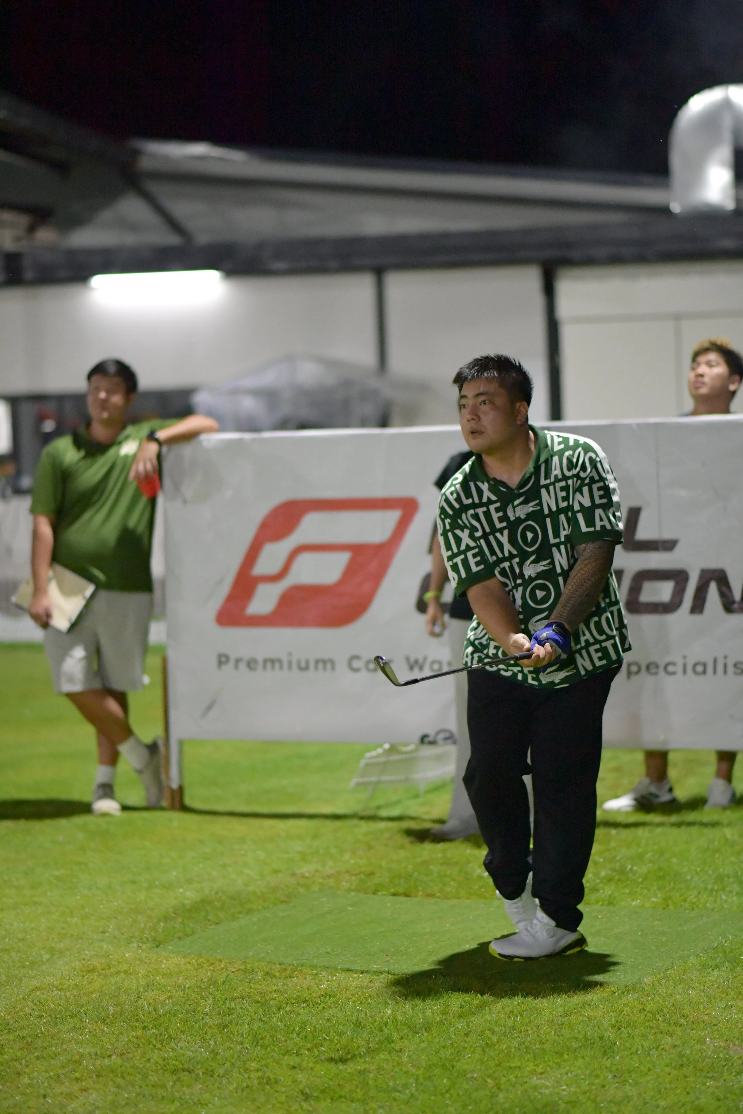 A man in a green and black shirt playing golf on a grassy field at night, with two other men standing behind a barrier watching.