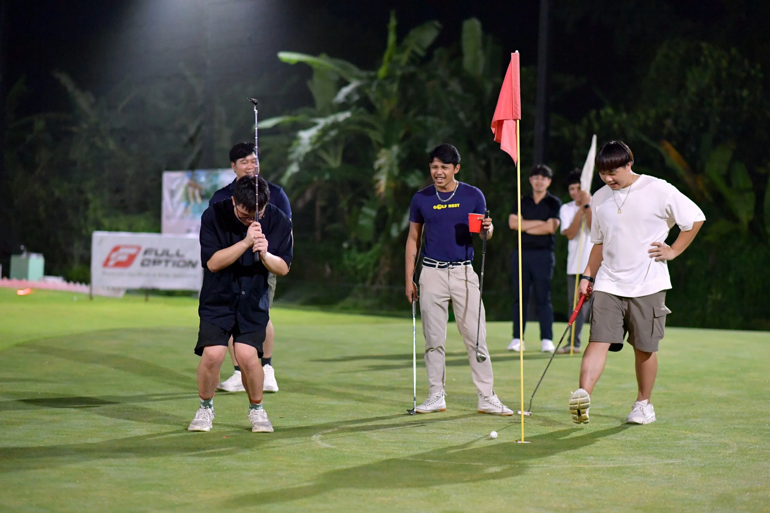 Group of friends playing golf on a putting green at night, some smiling and enjoying the game.