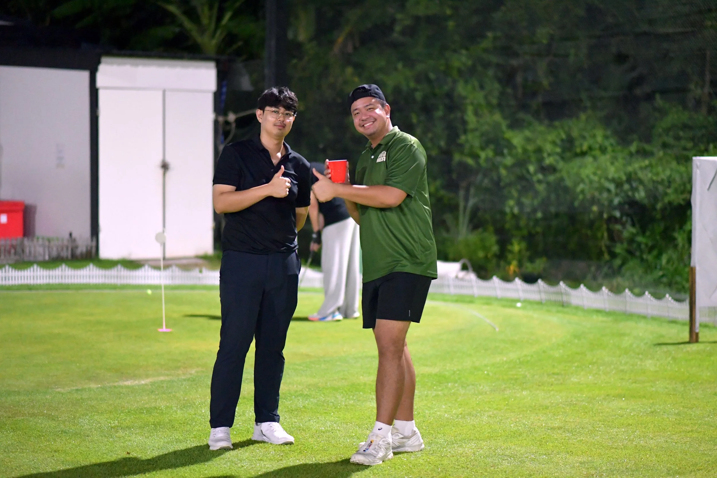 Two men standing on a well-lit golf course at night, one giving a thumbs up and the other smiling while holding a red mug, with greenery and golf equipment in the background.