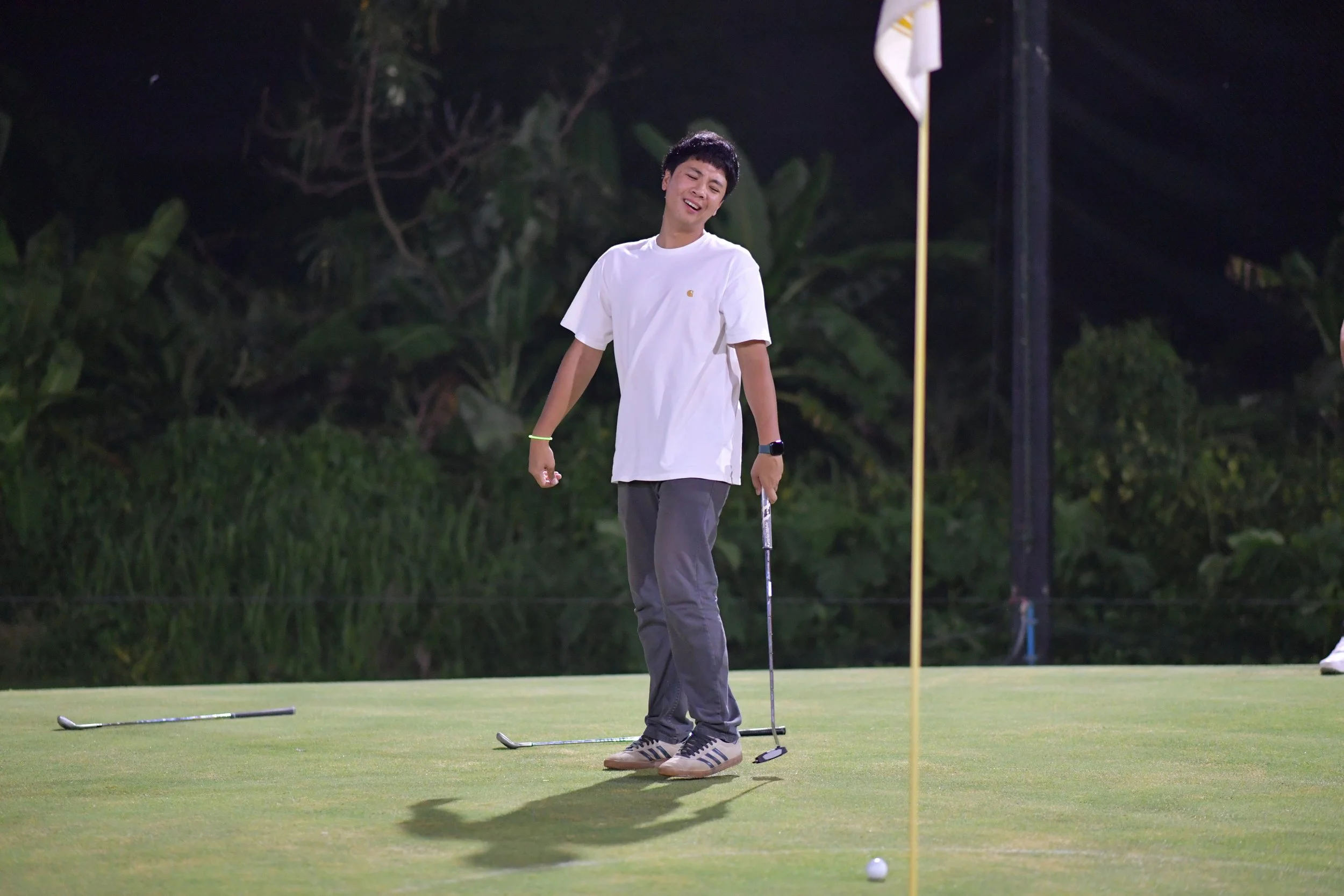 A young man smiling and holding a golf club stands on a putting green at night, with golf balls and flags nearby, surrounded by tall greenery.