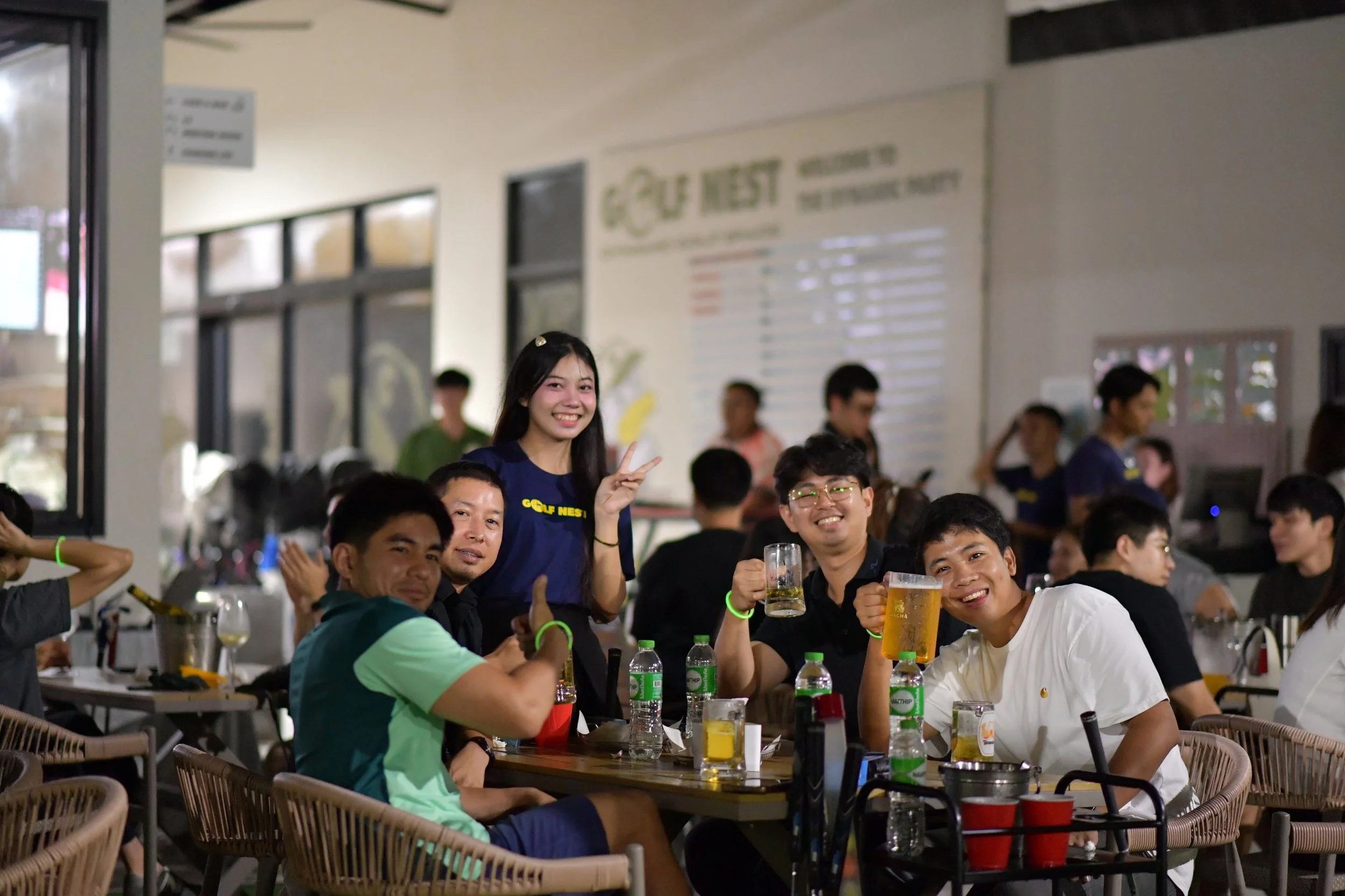 A group of five young people sitting and standing around a table at a restaurant or bar, smiling, holding drinks, and making gestures, with other people in the background.