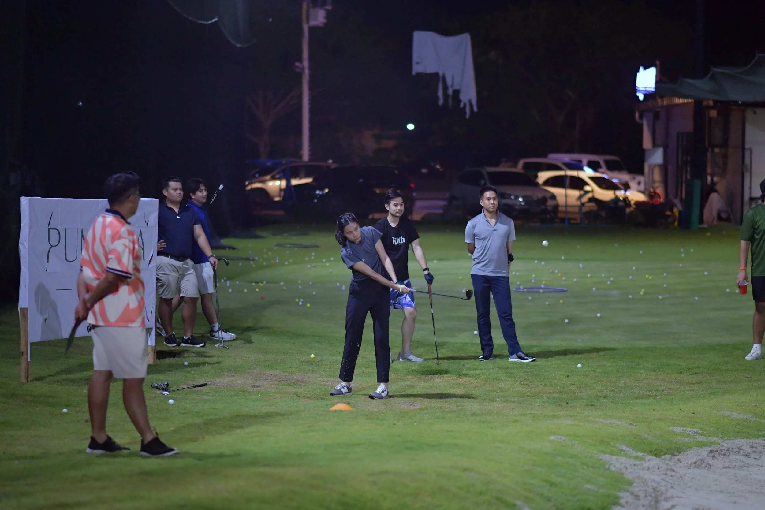 Group of people practicing golf on a driving range at night with scattered golf balls around.